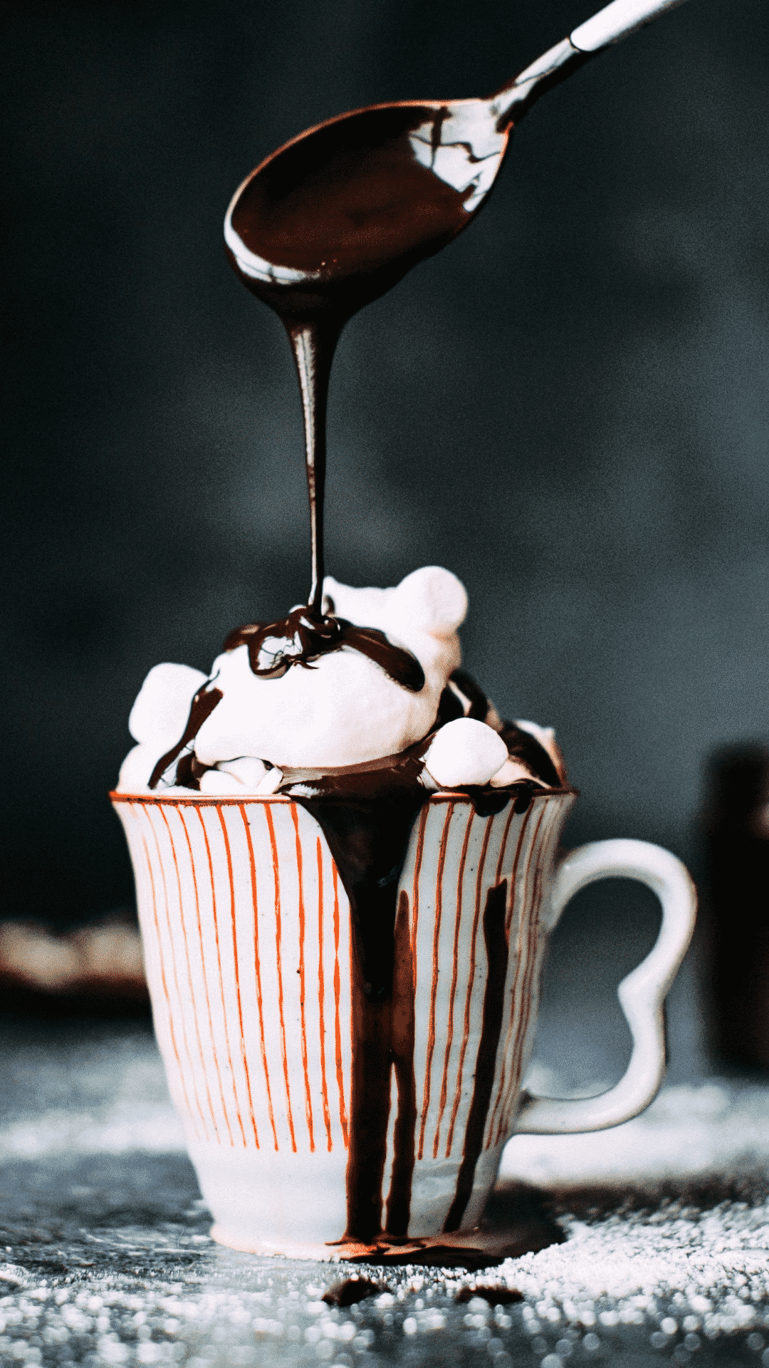 A cup of hot chocolate with marshmallows and chocolate sauce being poured over the top. - Marshmallows, chocolate