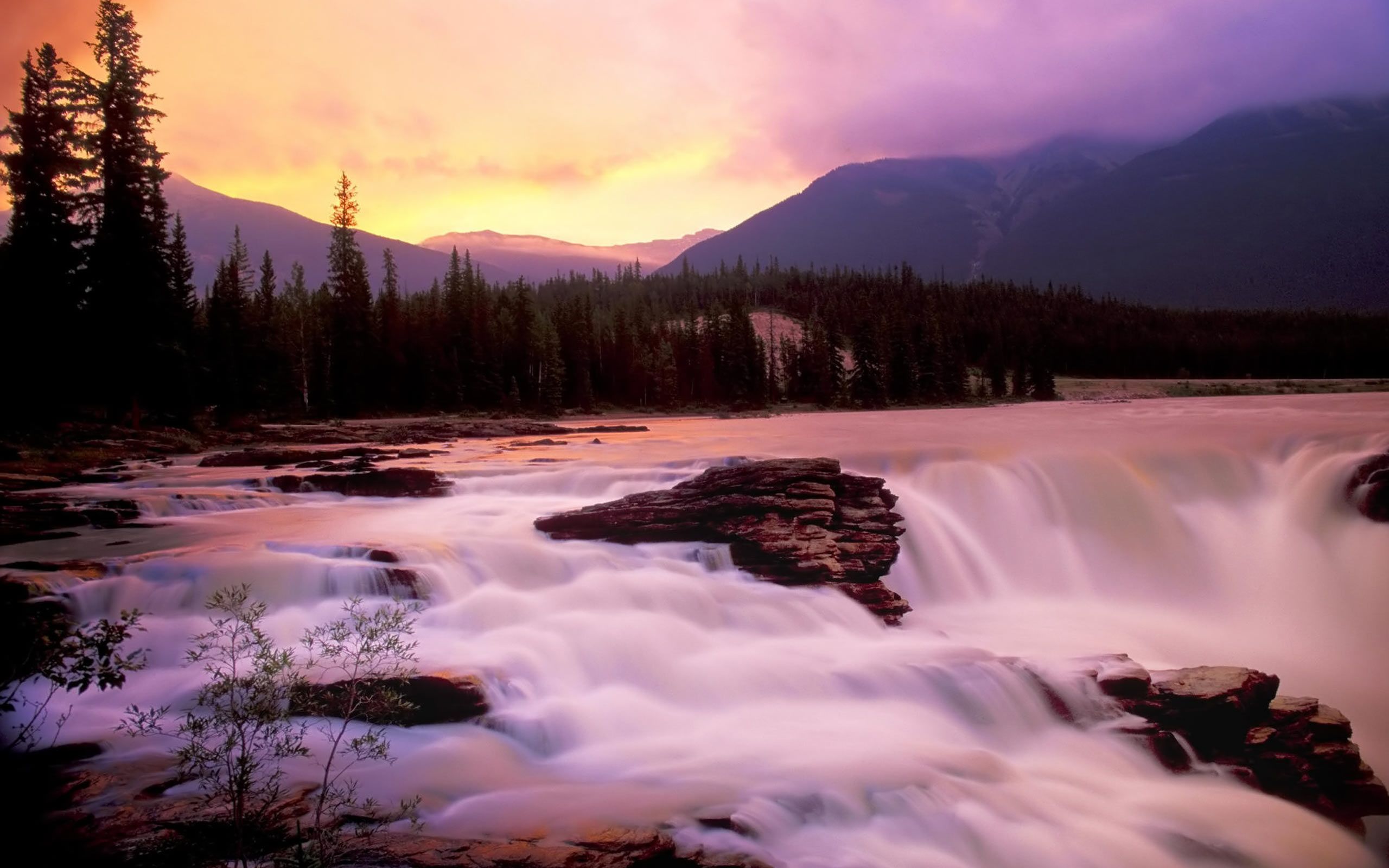 A river flows over rocks in front of a mountain at sunset. - Waterfall