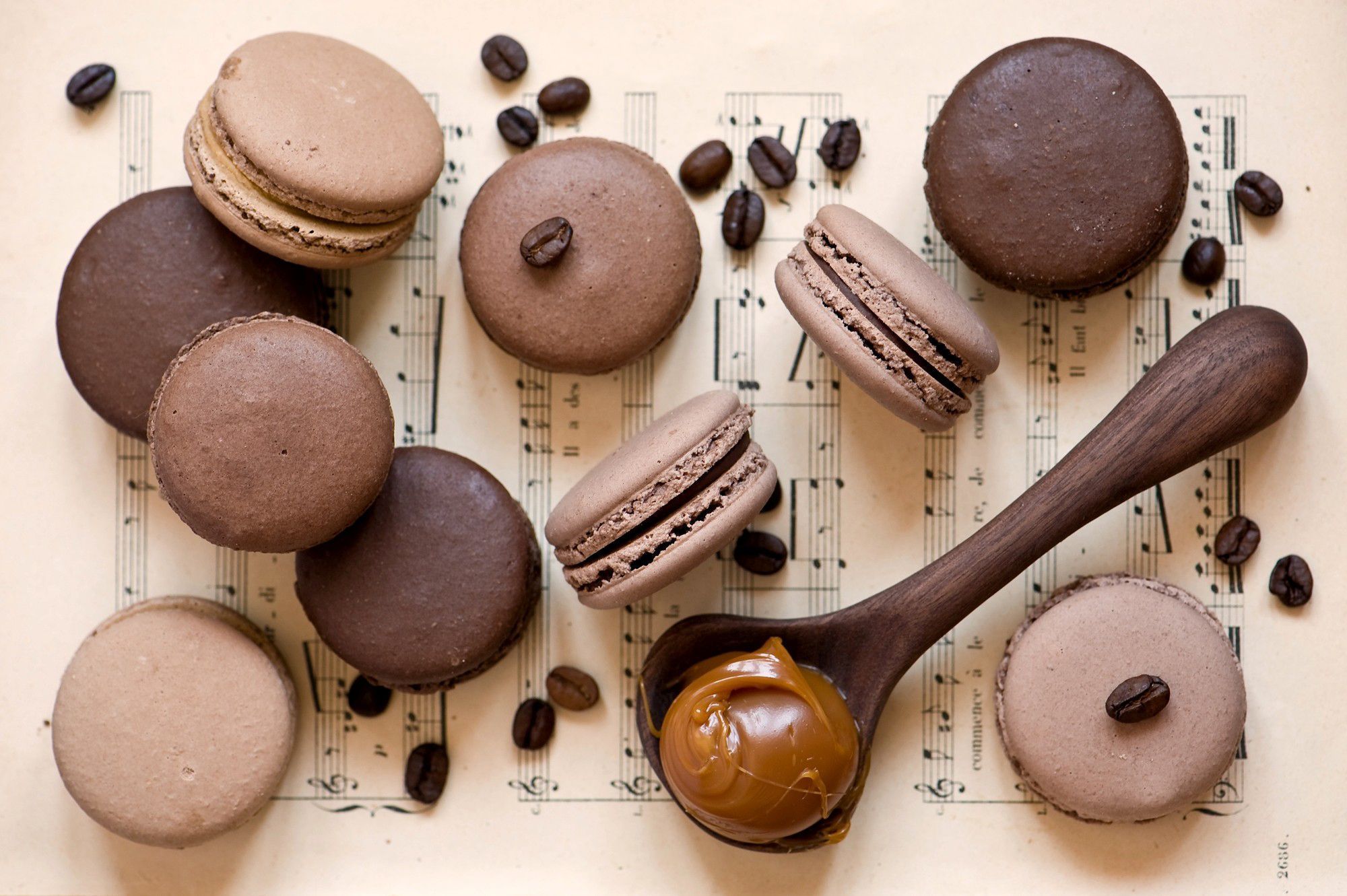 A wooden spoon with caramel on it is surrounded by chocolate macarons and coffee beans. - Macarons