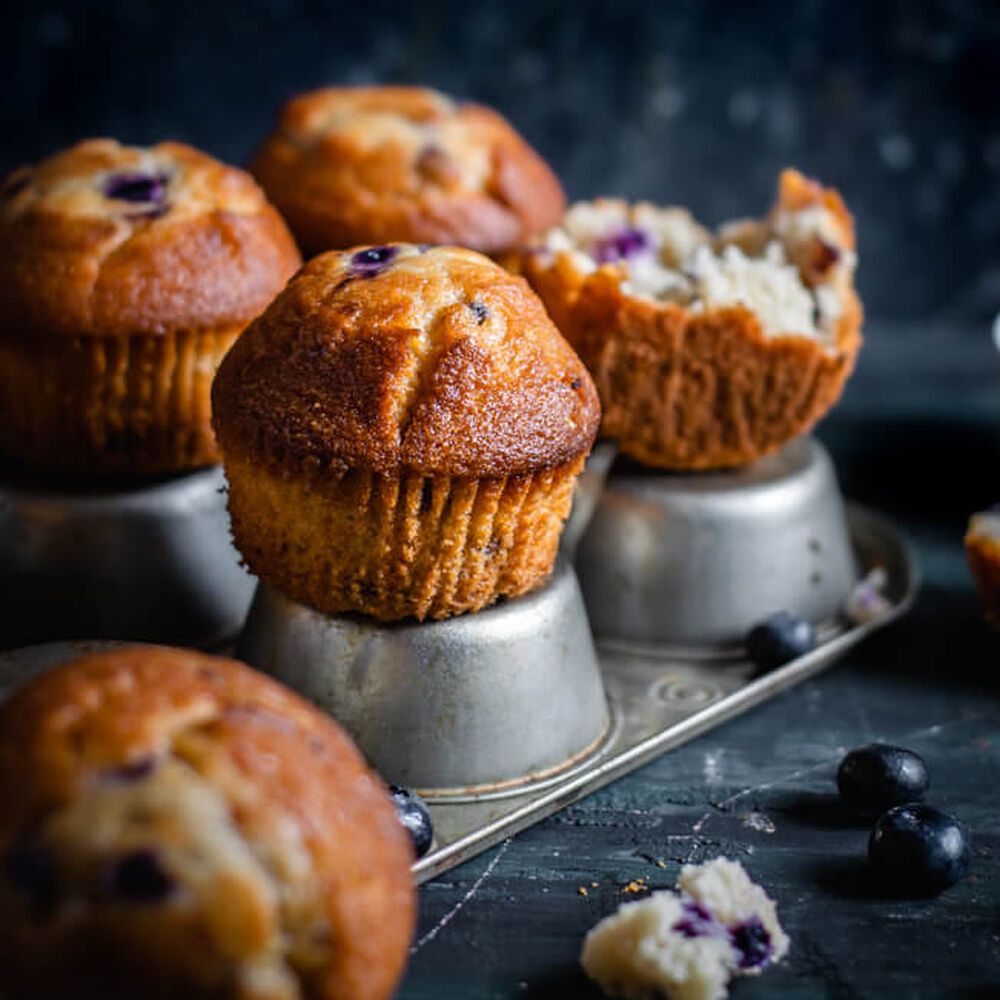 Blueberry muffins on a muffin tray with a couple of muffins in the foreground and a blue background. - 