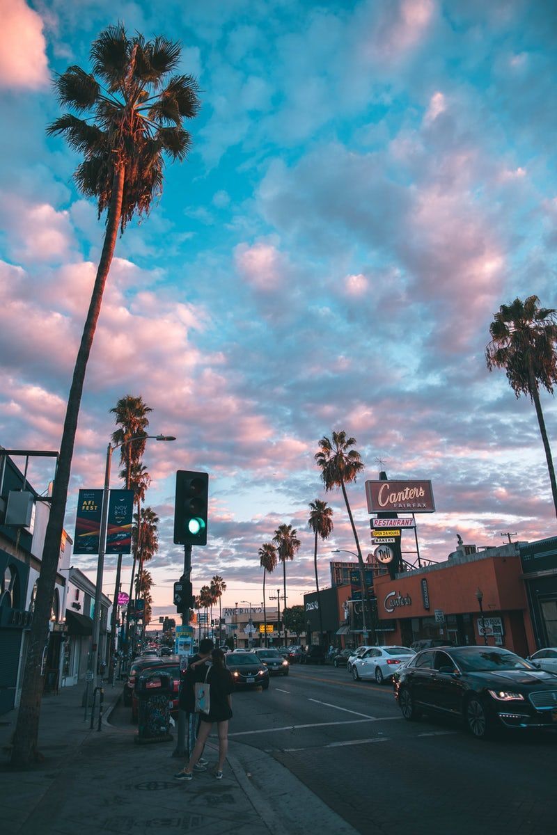 A sunset view of a street with palm trees and a couple waiting to cross the street. - California