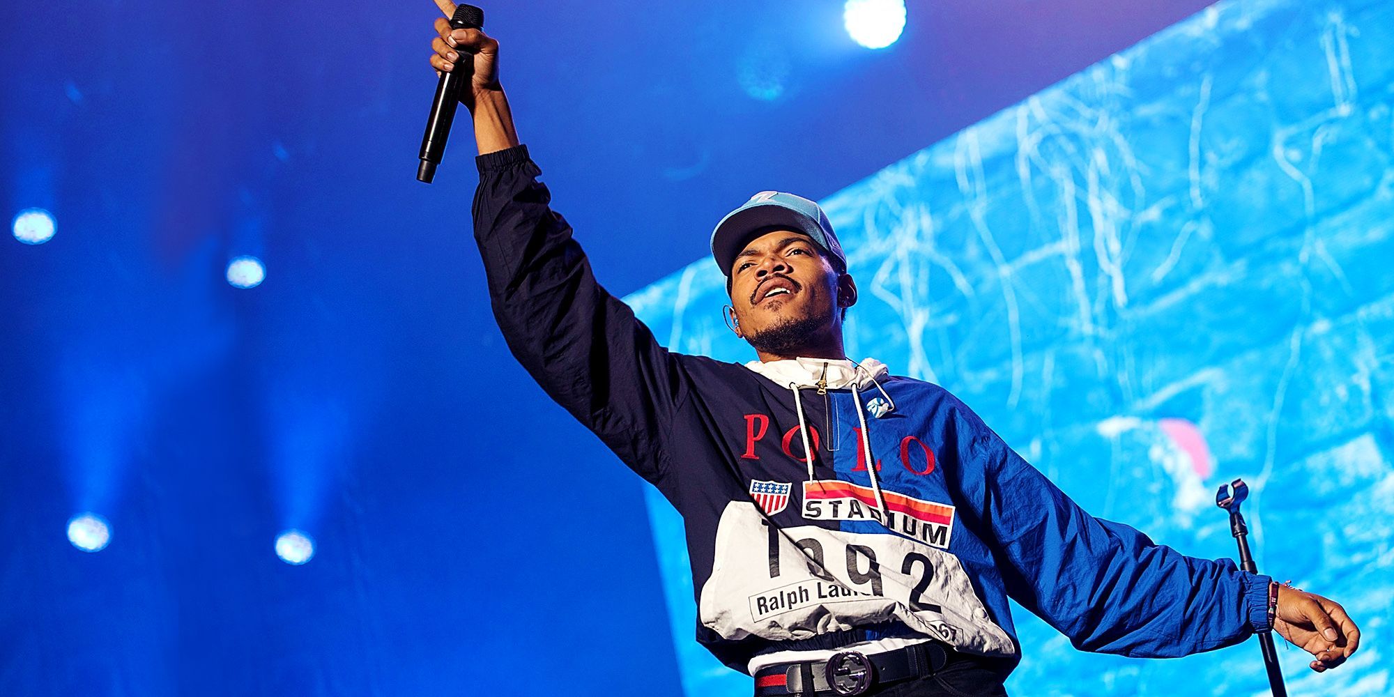 A man in blue jacket and hat holding up his hand - Chance the Rapper