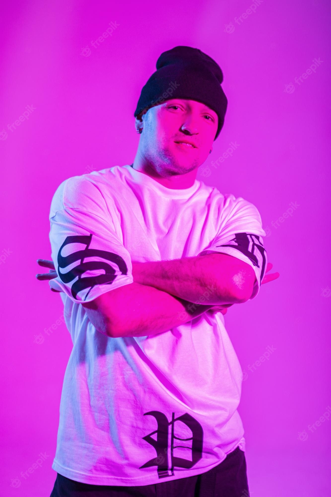 A man in a white t-shirt and black beanie stands with his arms crossed in front of a purple background. - Chance the Rapper