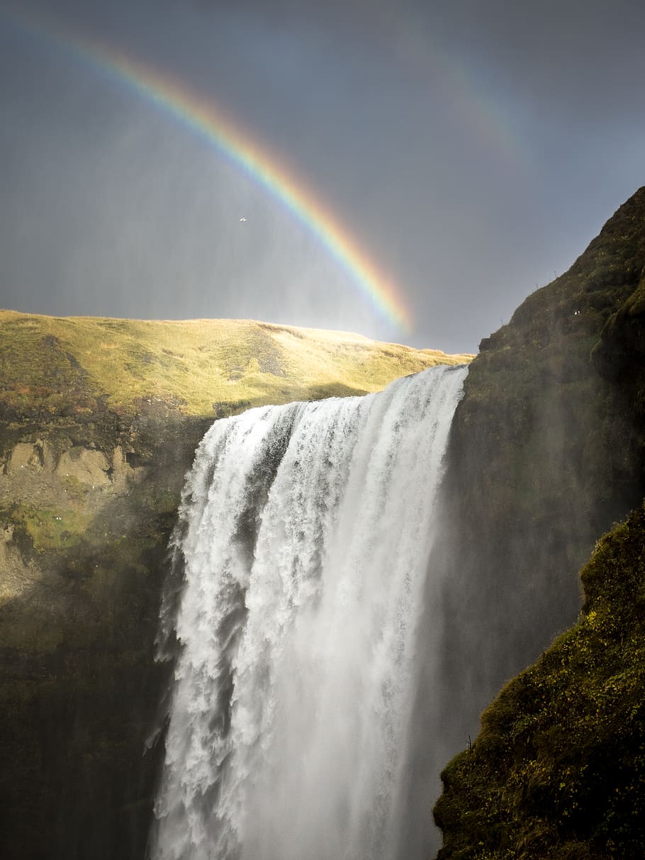 This image features a beautiful waterfall cascading down into a river, with a rainbow visible behind the waterfall on the left side. The rainbow appears to be in the shape of a double rainbow, with multiple shades of color. The waterfall is surrounded by various trees, including a dense section of trees in the upper left corner of the image. There are several people scattered throughout the image, with some  - Waterfall