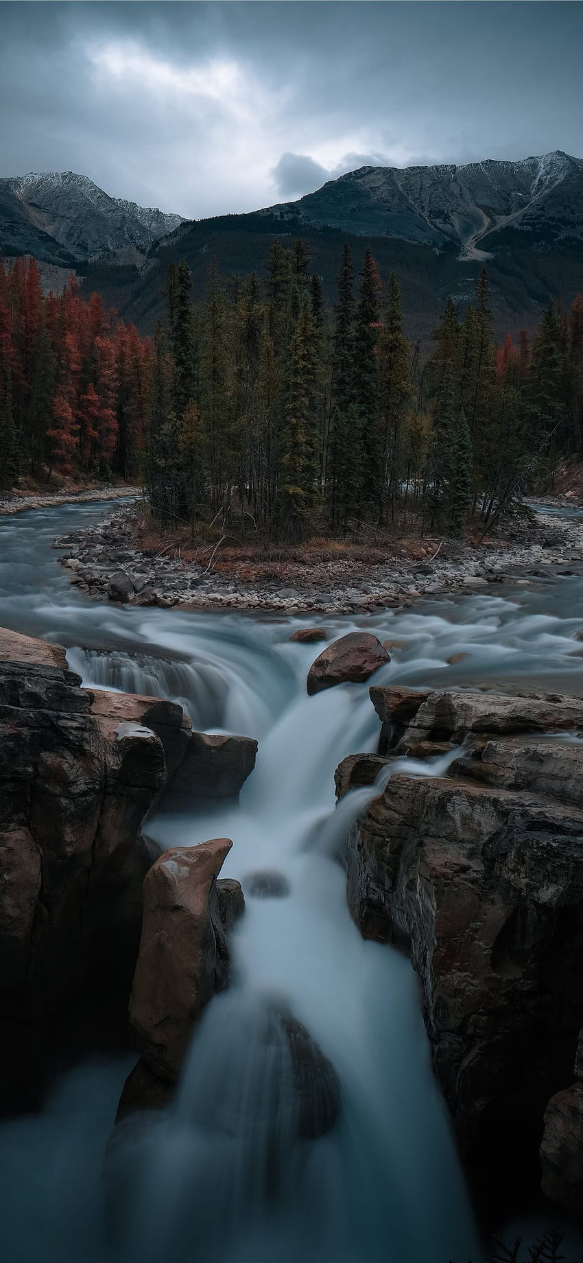 A stunning landscape is featured in this image, featuring a fast flowing river with rocks in its path. The river is surrounded by trees and mountains, creating a picturesque scene. The flowing water is divided into numerous streams, each showcasing an intense force of  - Waterfall