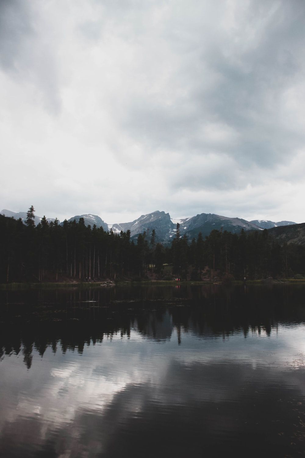A cloudy sky reflects in a lake surrounded by trees and mountains in the background. - Lake