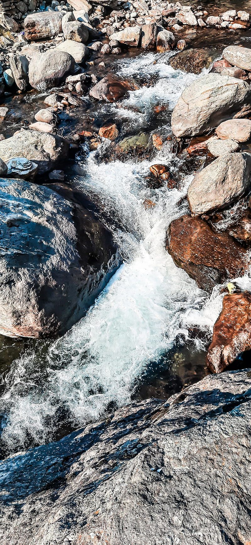 A scene of a mountain stream is portrayed, with water flowing over rock formations in the background. A hiker is seen standing near the water, enjoying the natural beauty of the stream. There are several rocks scattered around the stream, providing various places for the hiker to rest, take pictures, or simply enjoy the view. In the foreground, a rock is positioned closer to the water, partially submerged and creating a small, shallow pool. Another rock is visible on the right side of the picture, serving as a point of interest for the hiker. The hiker is holding a pair - River