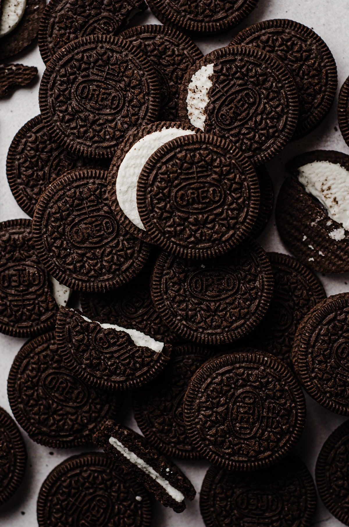 A photograph captures a large pile of Chocolate Chip Oreo cookies on a white surface. The cookies are arranged in a cluster with several of them already broken into smaller portions or completely consumed. Some of the cookies are partially crushed or crumbled, indicating that they have been eaten or attempted to be eaten. The pile of cookies is quite large, and it appears that there are several cookies scattered around the surface. Many of the cookies are visible and easily distinguishable, while some are partially covered by other cookies or located near the edges of the image. The overall image showcases a - Oreo