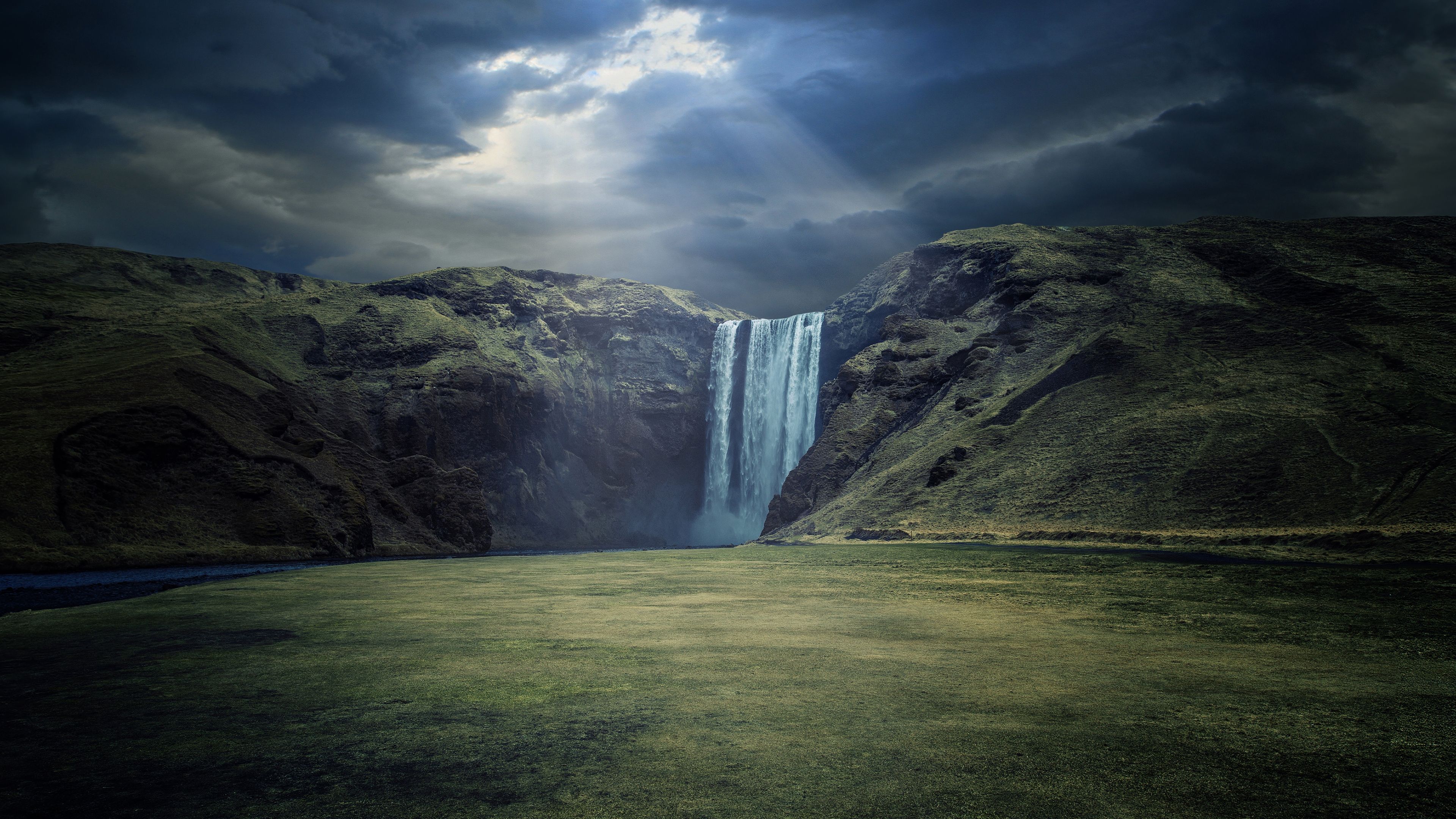 A waterfall cascades down a grassy hillside under a cloudy sky. - Waterfall