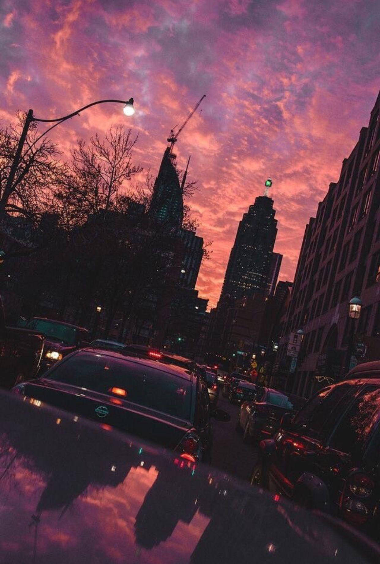 A city street with cars and tall buildings with a pink and purple sunset in the background. - Sunset