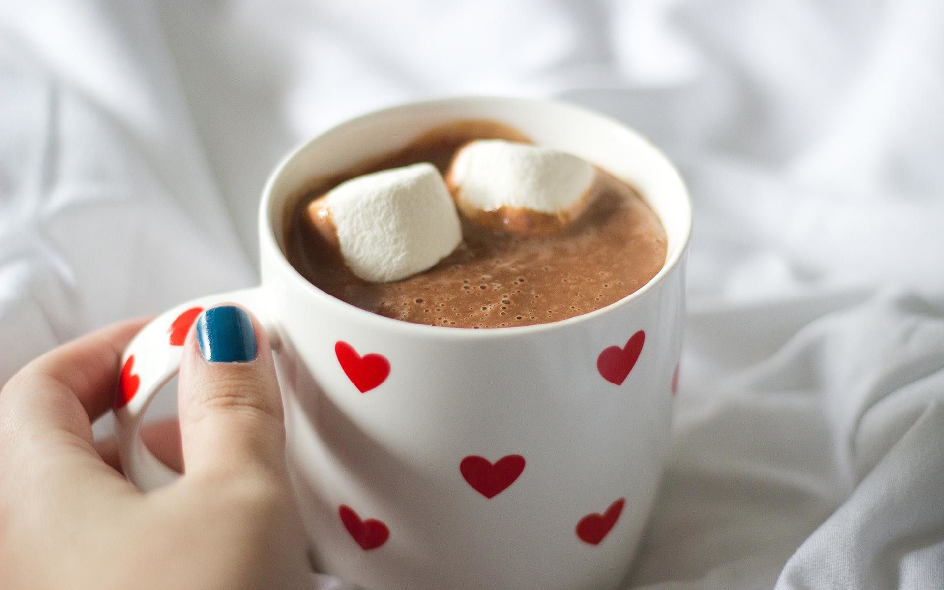 A woman's hand holding a white mug with red hearts on it, filled with hot chocolate and marshmallows. - Marshmallows