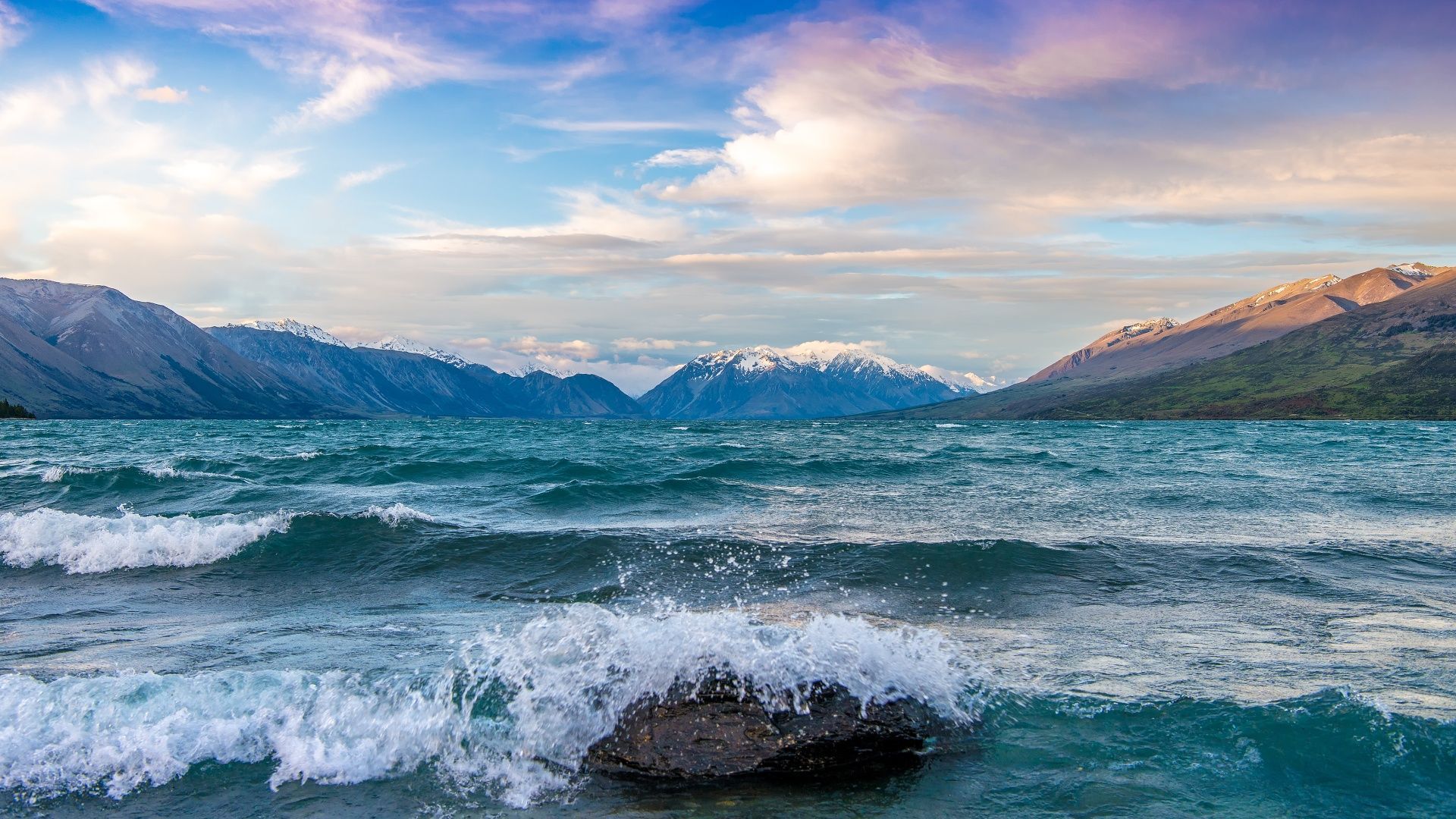 The ocean waves crashing against a rock in the foreground with snow capped mountains in the background. - Lake