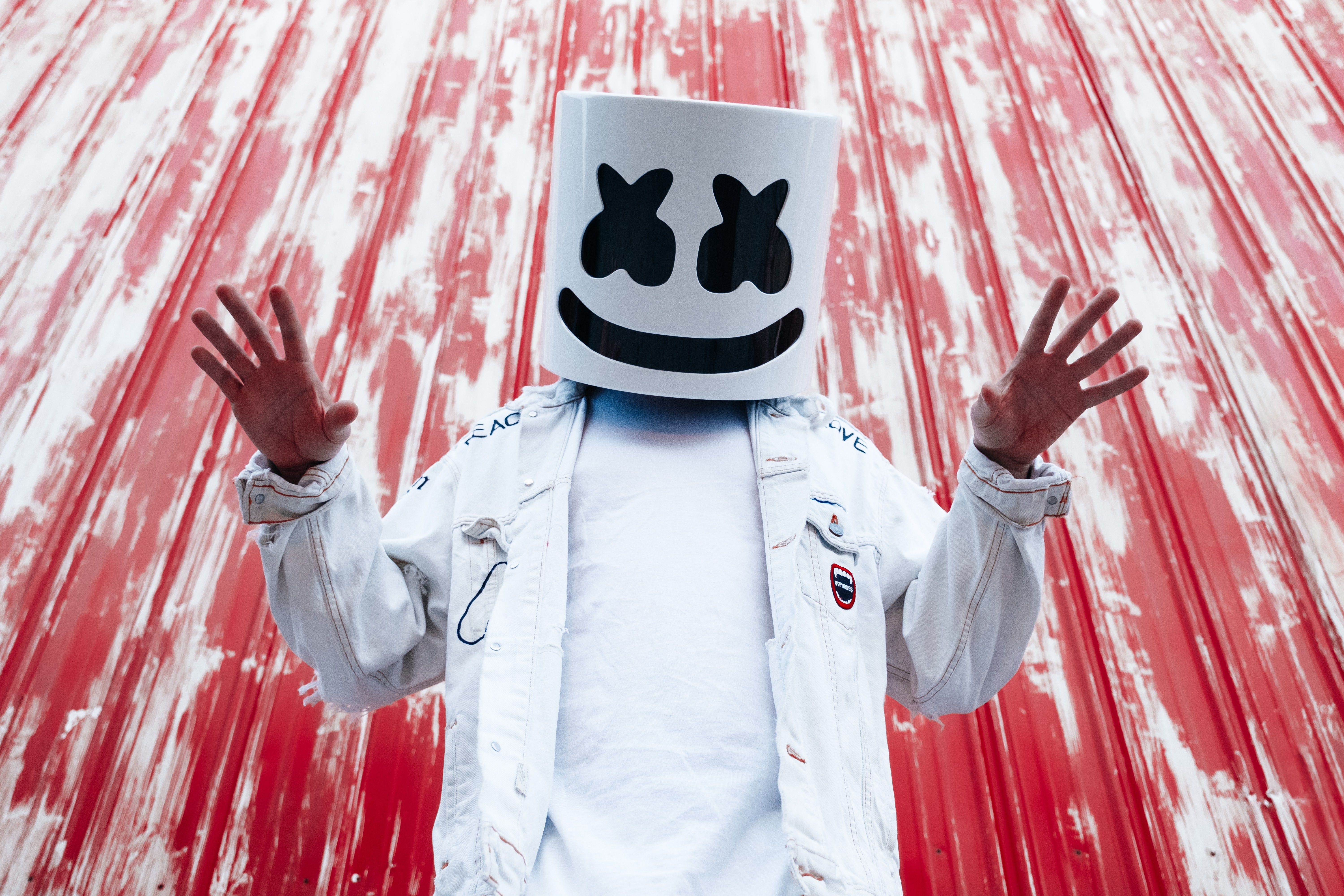 A man wearing a marshmallow headpiece and a white jacket in front of a red background - Marshmello