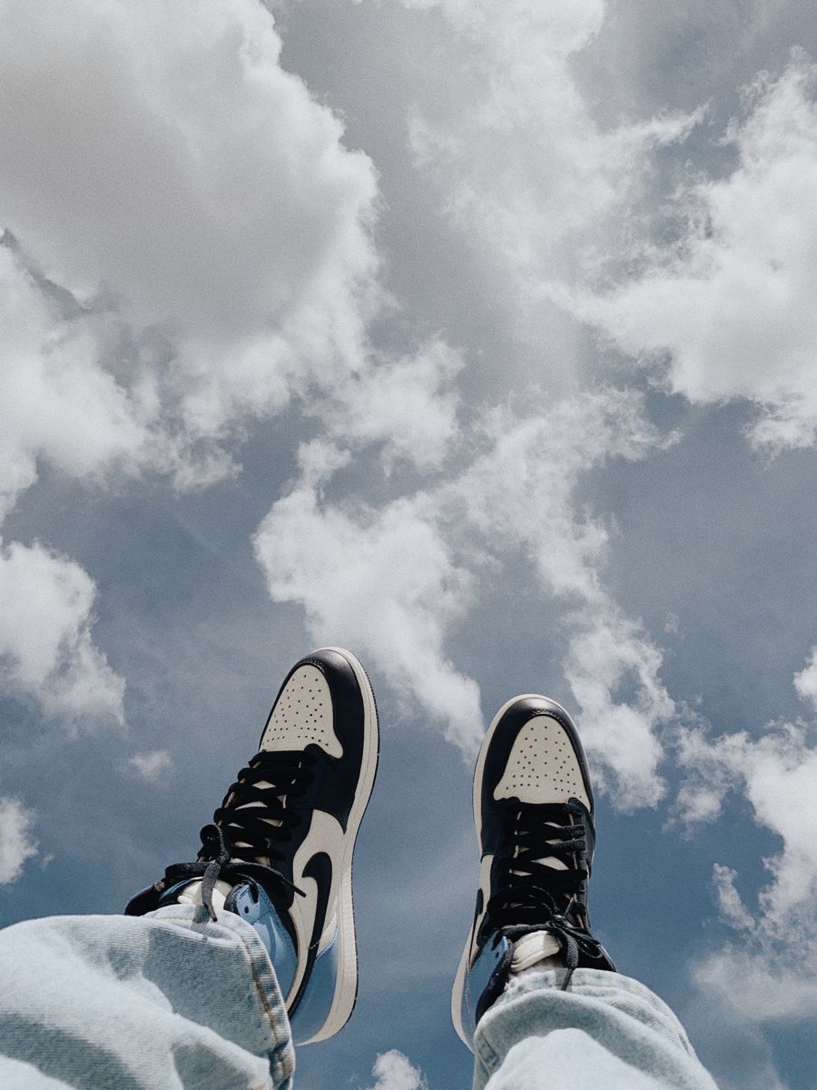 A person's feet wearing black and white Nike sneakers and jeans are shown against a blue sky with white clouds. - Air Jordan