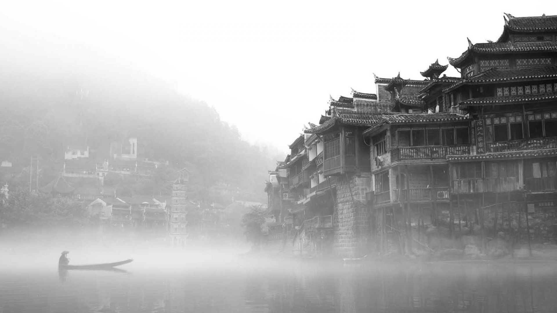 A black and white photo of a man rowing a boat in a river in front of a traditional Chinese village. - Chinese