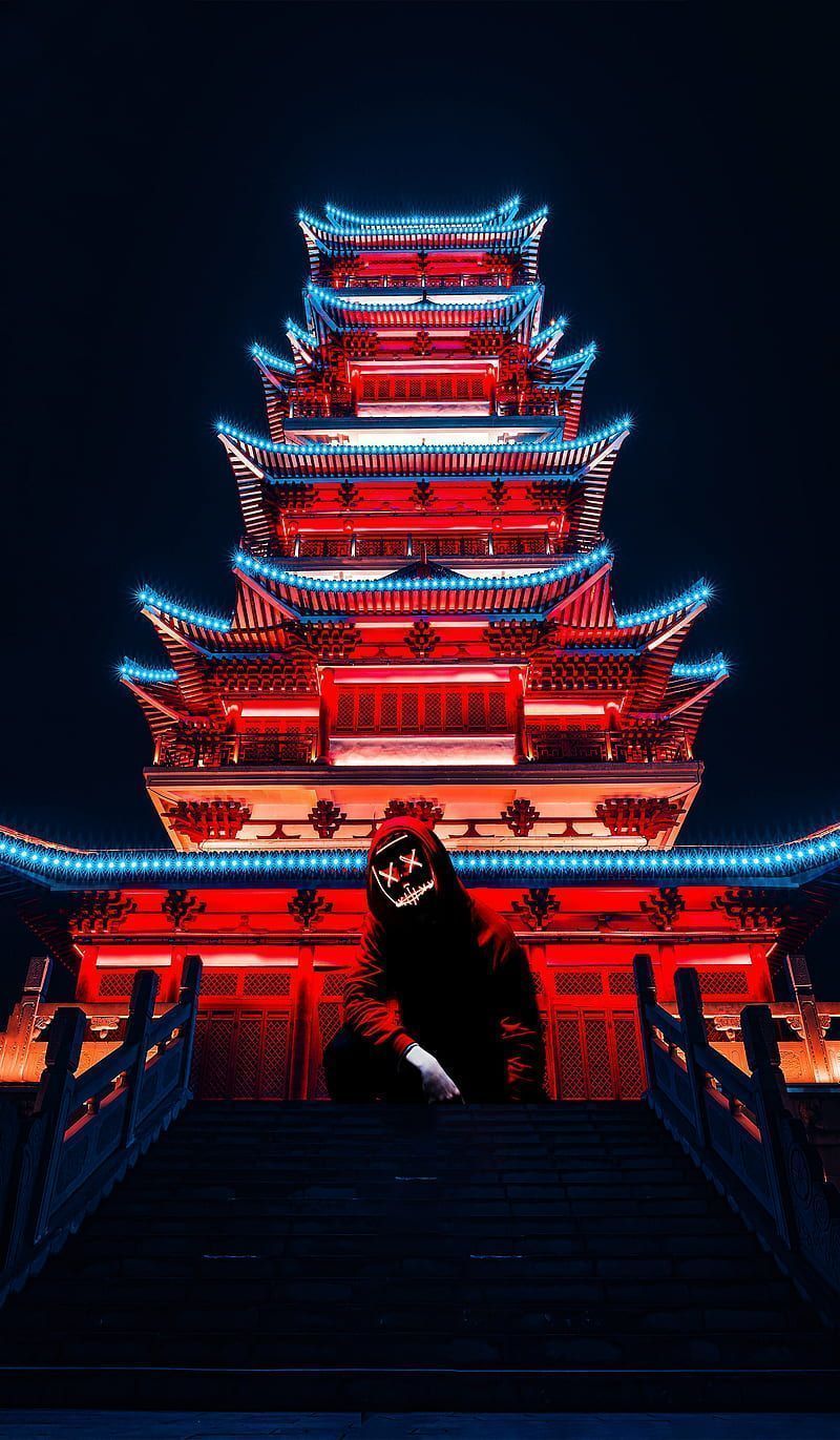 A person in a black hoodie with a white face mask sitting in front of a red lit pagoda - Chinese