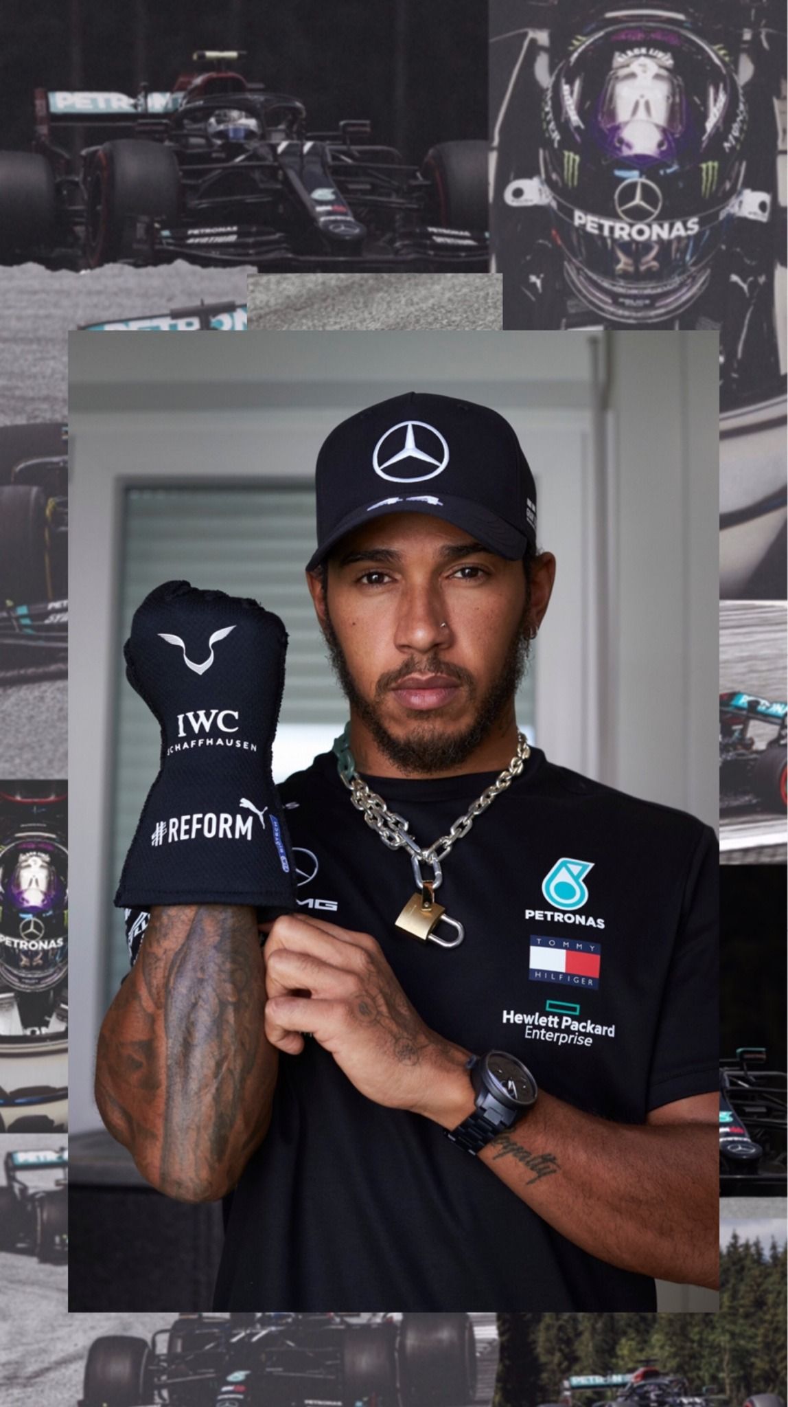 Lewis Hamilton, wearing a black Mercedes cap and t-shirt, holds a black IWC cap in front of a wall of Mercedes cars. - Lewis Hamilton