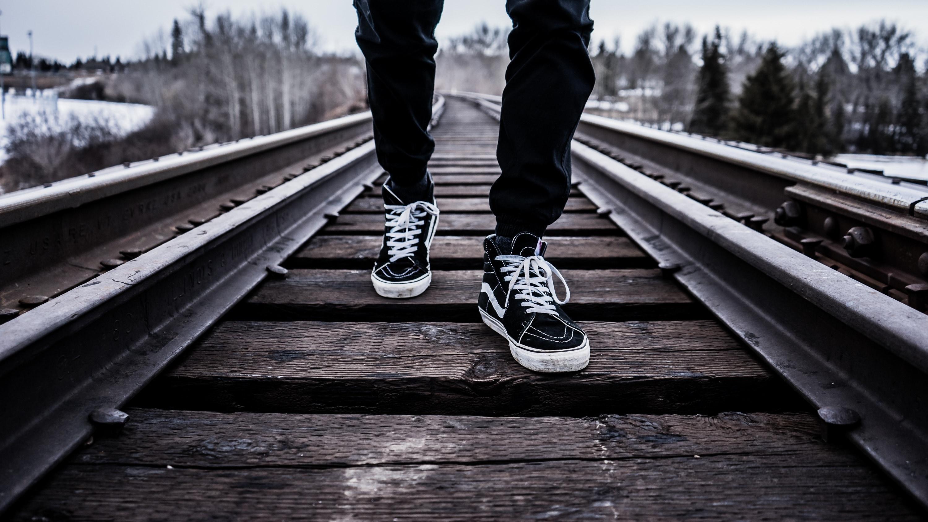 A person wearing black pants and black and white sneakers is walking on the train tracks. - Vans