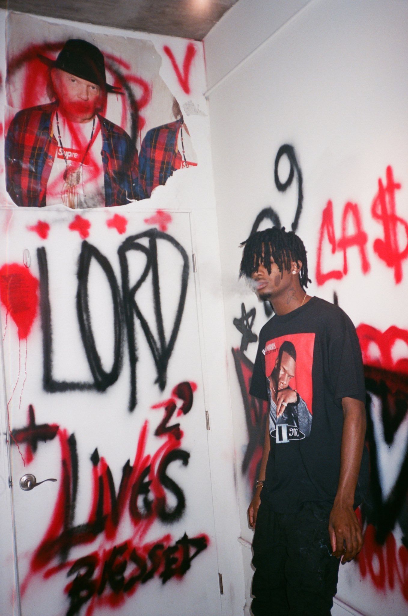 Travis Scott wearing a black graphic tee with a red box logo and standing in front of a graffitied wall - Graffiti