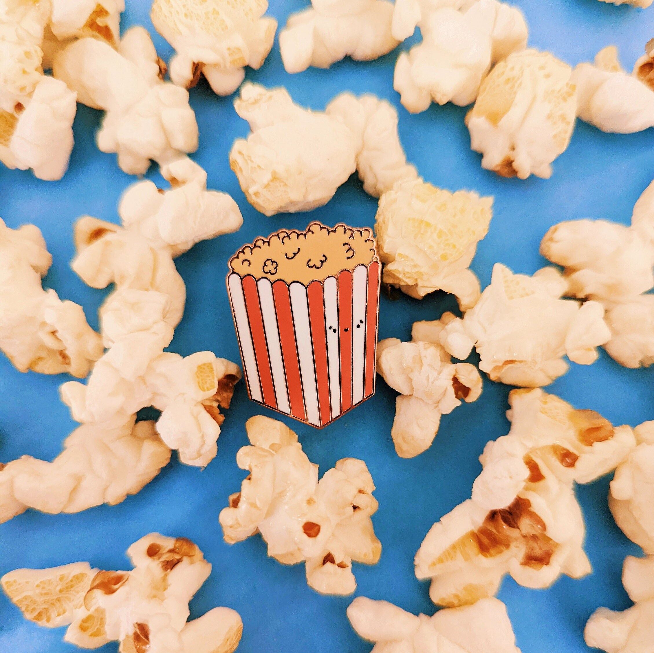 A hard enamel pin of a kawaii popcorn box, with the popcorn flying out of the box. - Popcorn