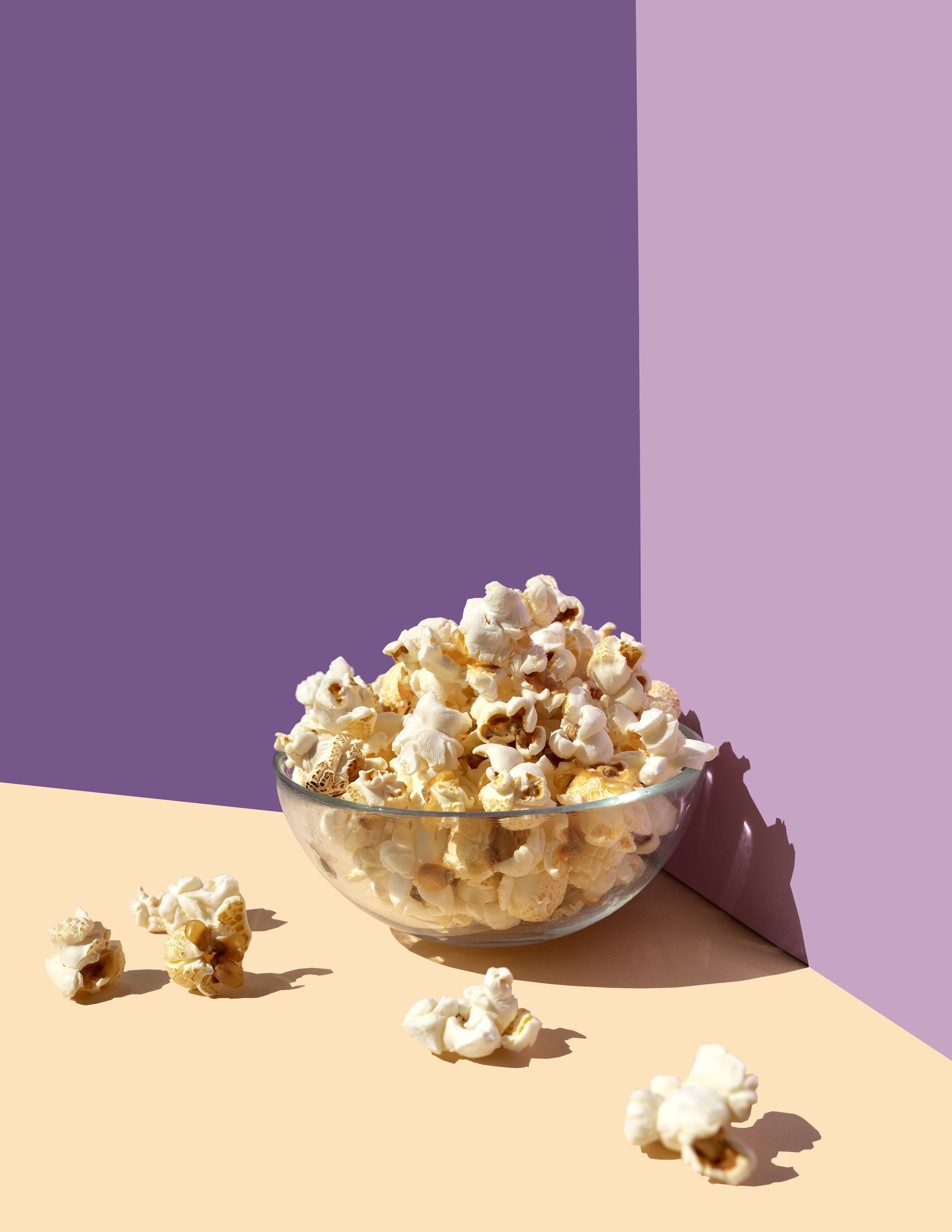 A bowl of popcorn sits on a table with a purple and pink background. - Popcorn