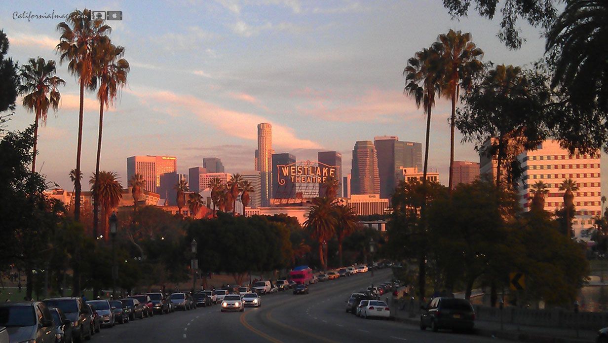 The sun sets over the city of Los Angeles, casting a warm glow over the buildings and palm trees. - Los Angeles