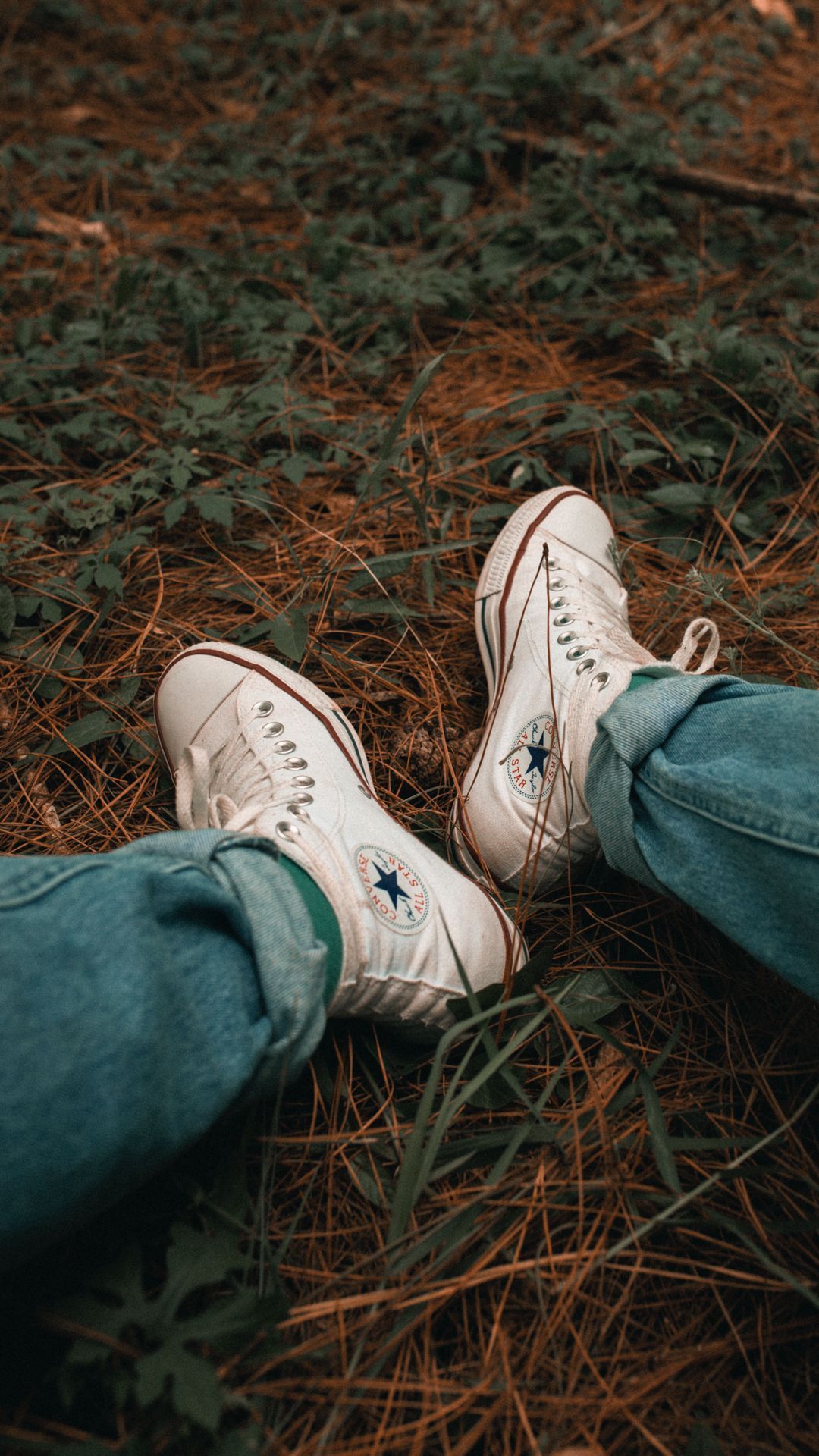 A person wearing jeans and white converse sneakers sitting on the grass - Converse