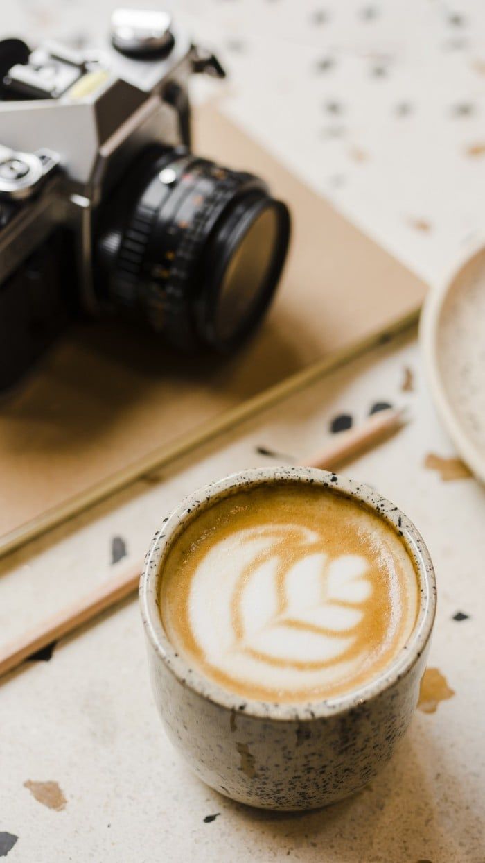 A cup of coffee with a leaf pattern in the foam, placed on a desk with a camera, a notebook and a pencil. - Coffee, warm