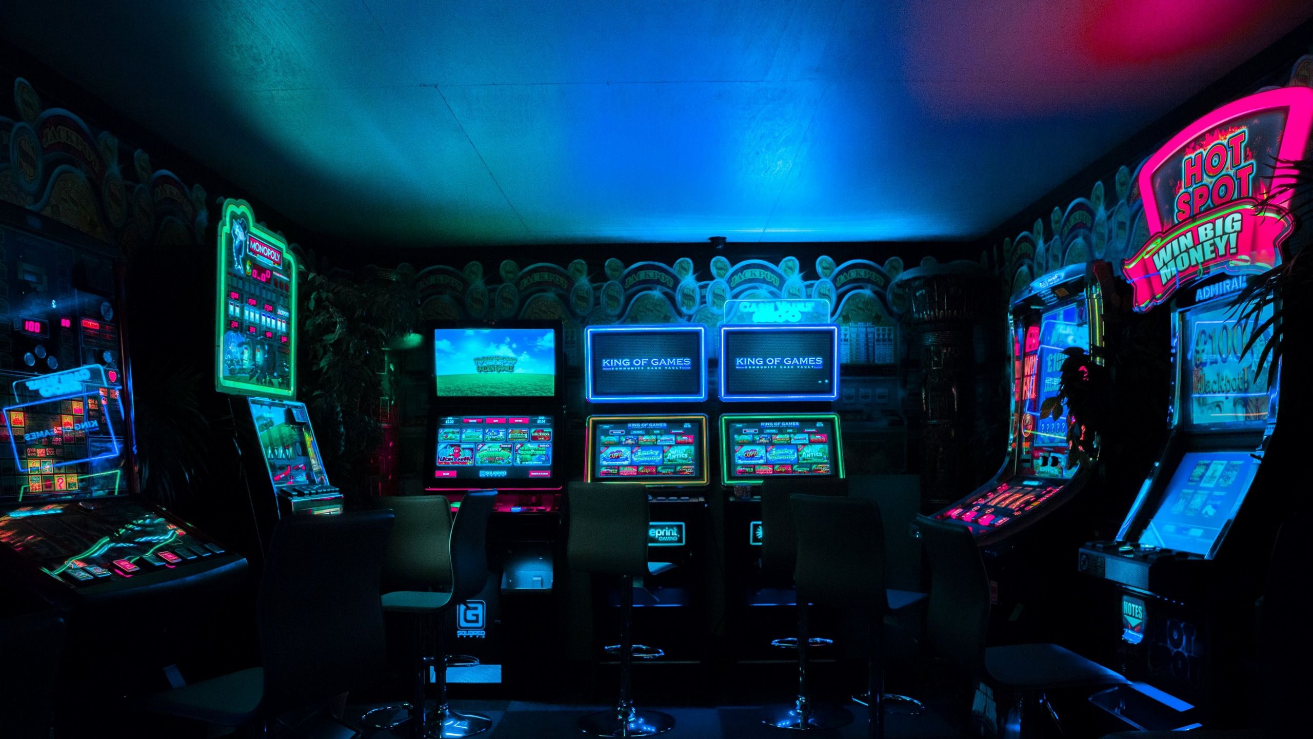 A dark room with multiple slot machines lit up by blue and pink lights. - Arcade