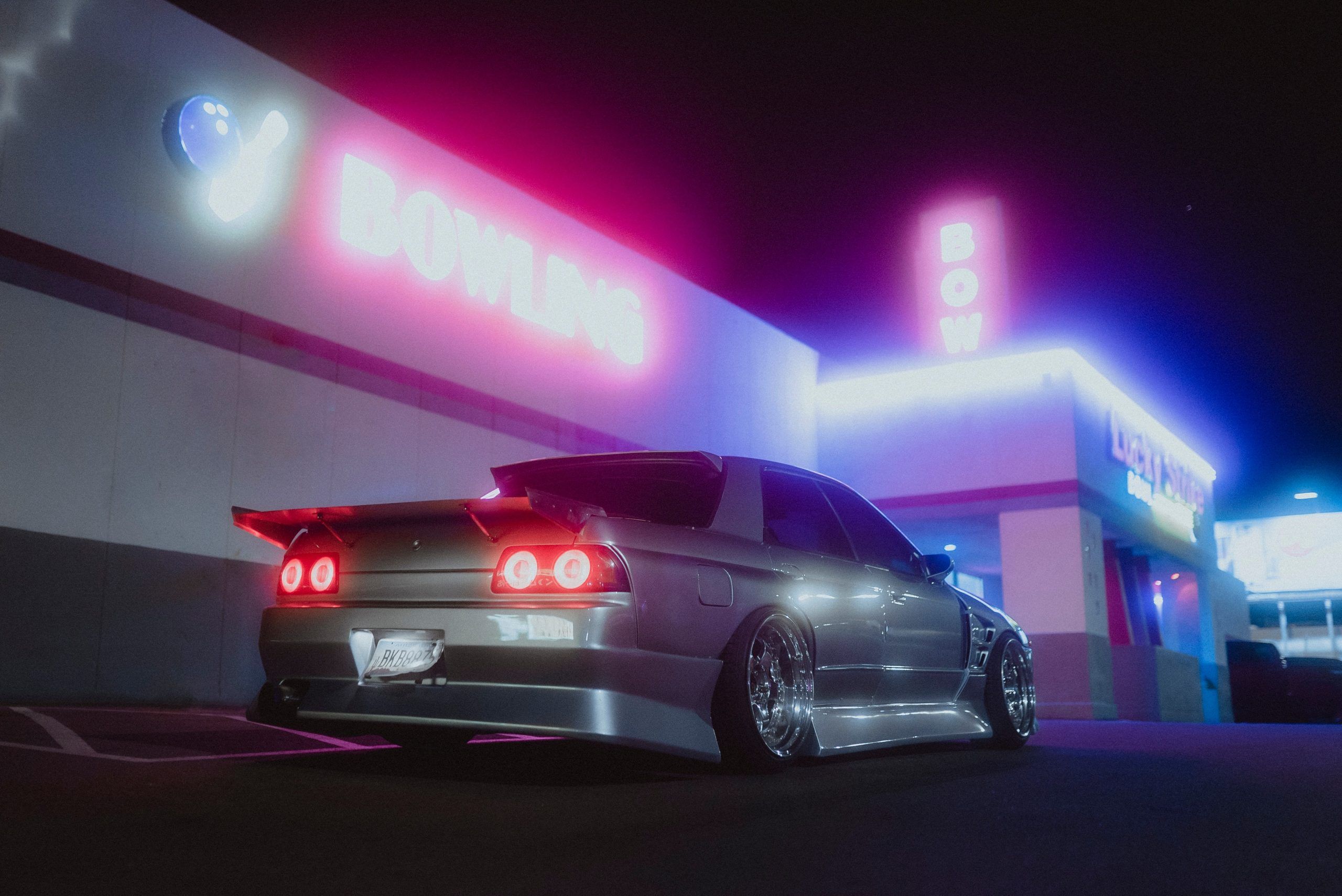 A grey car is parked outside a building with pink and blue neon lights. - Nissan Skyline