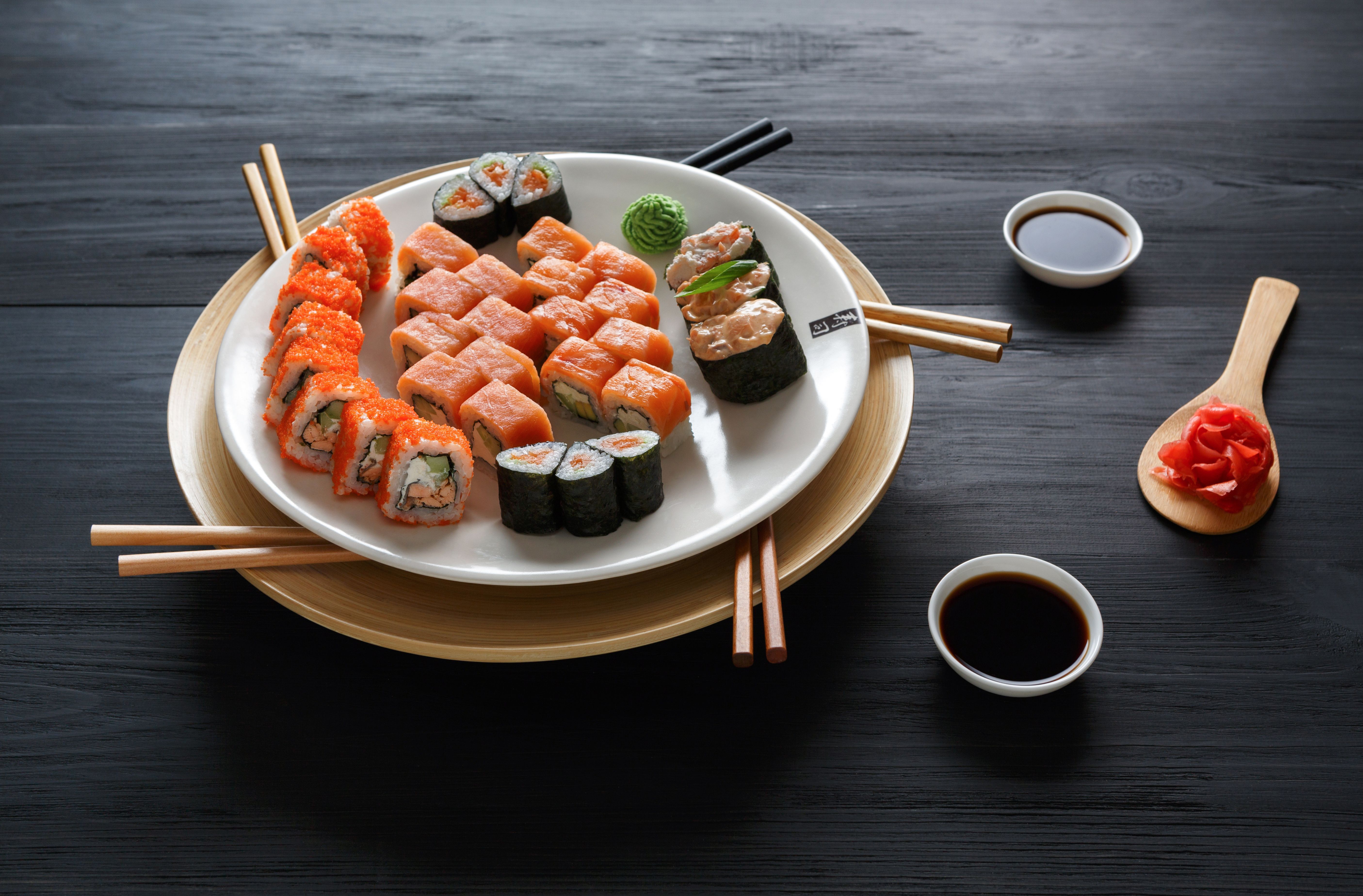 A plate of assorted sushi rolls with chopsticks and soy sauce on a black wooden table. - Sushi