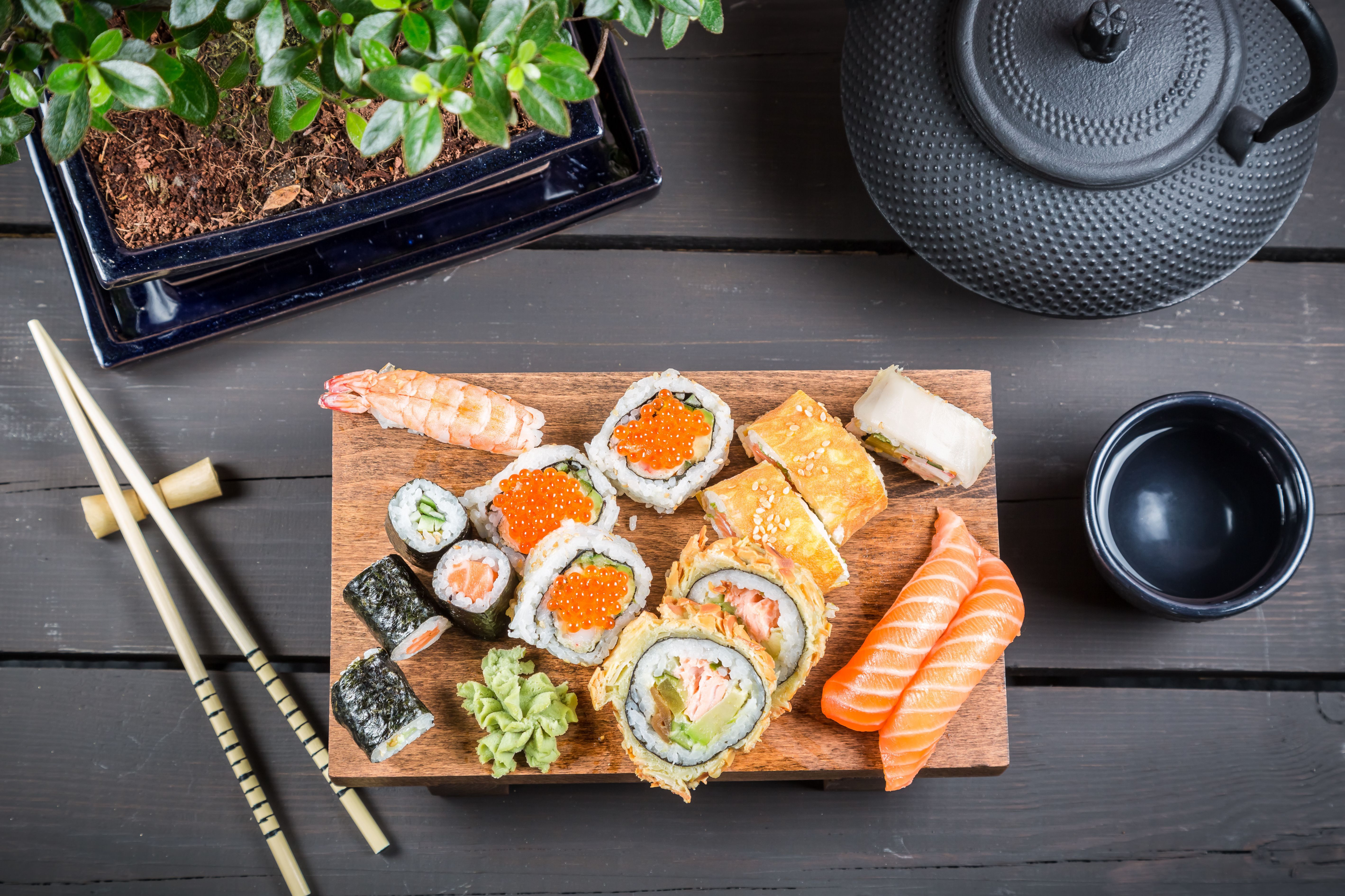 A variety of sushi and maki on a wooden board with chopsticks, a tea set, and a plant. - Sushi