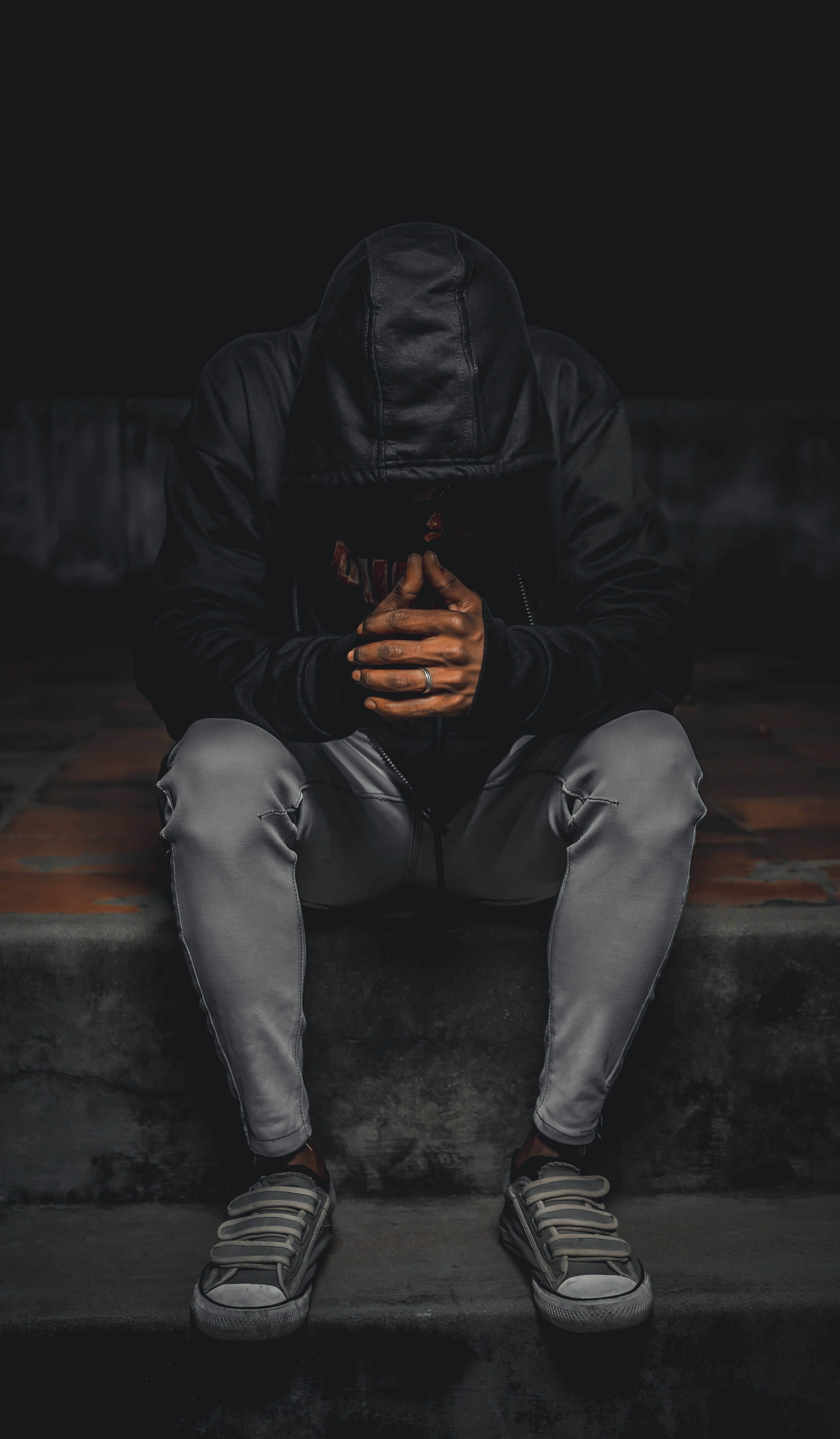 A man in a black hoodie sits on a step with his hands covering his face. - Depression