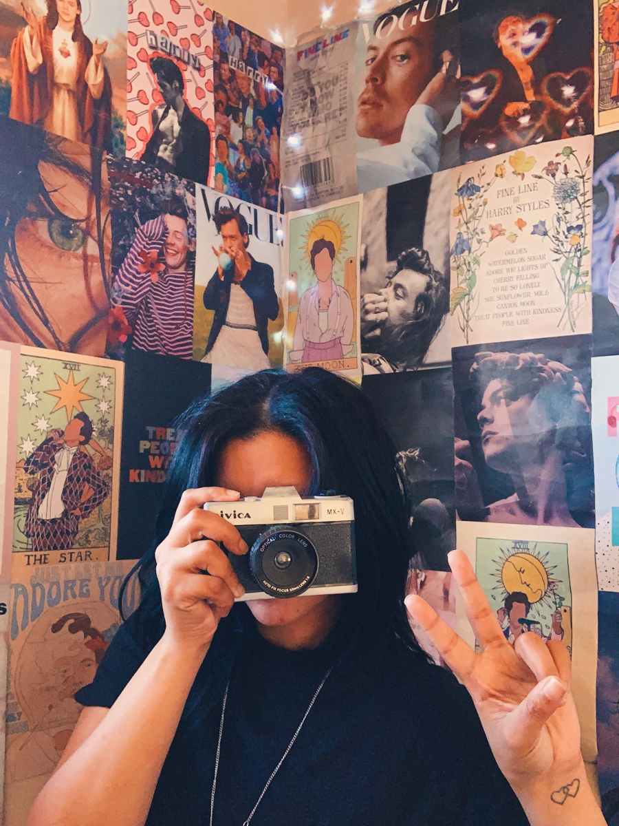 A girl with blue hair takes a picture in front of a wall of posters. - Harry Styles