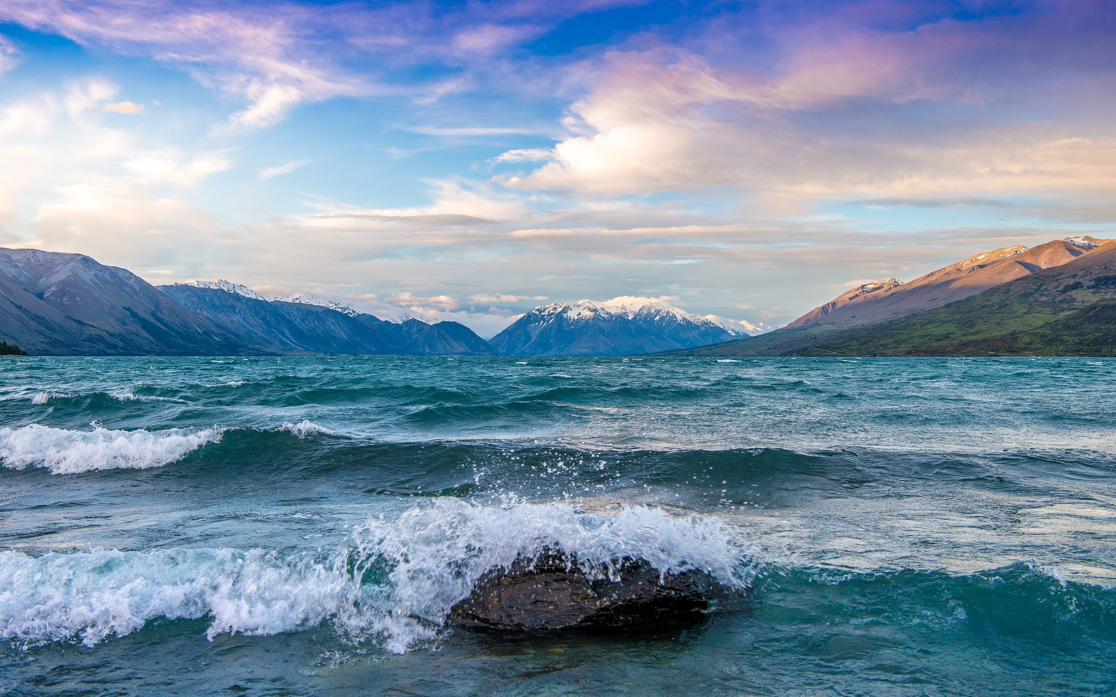 A wave crashes against a rock in the foreground of a mountain lake. - Lake