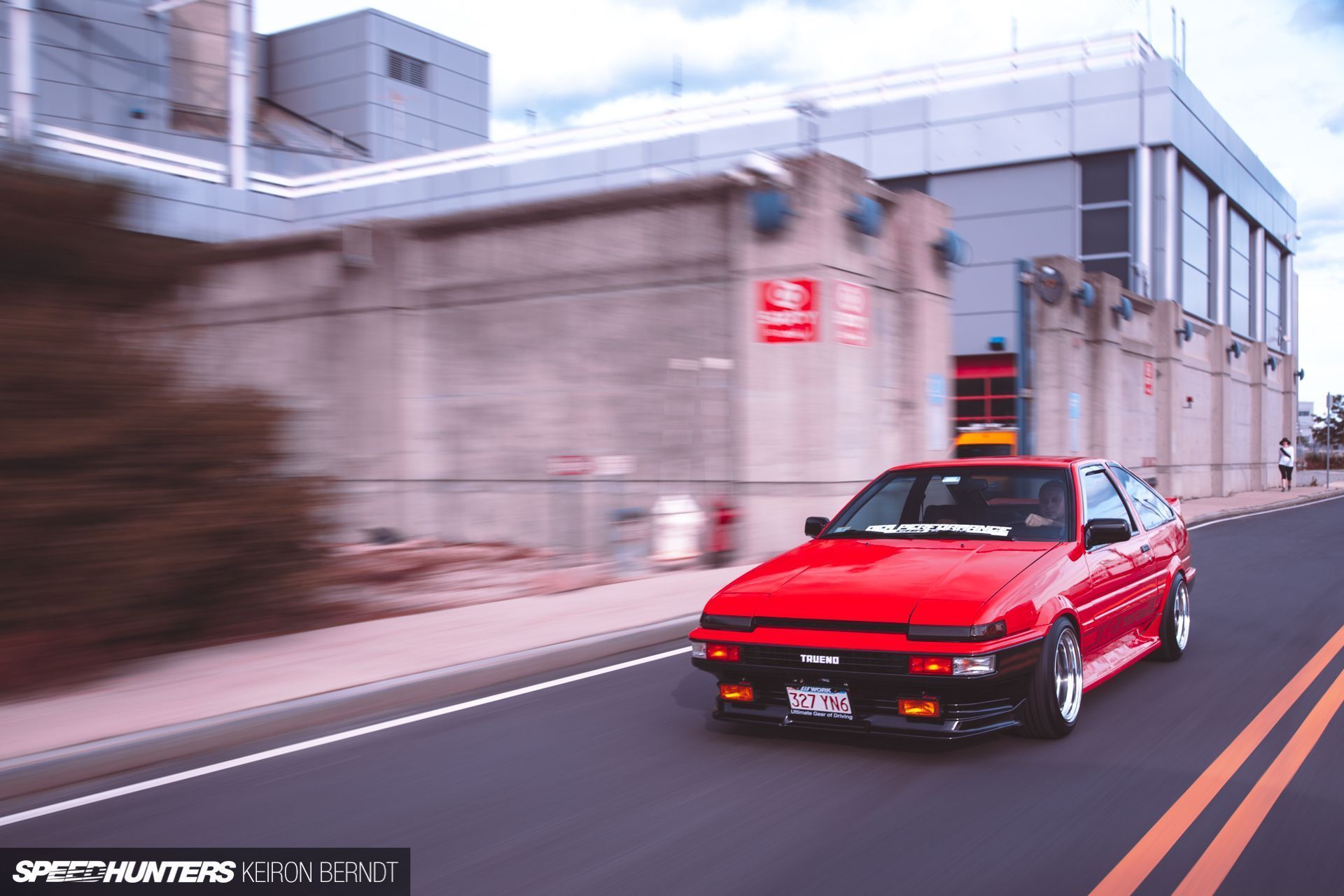 A red car driving down a street next to a building. - Toyota AE86
