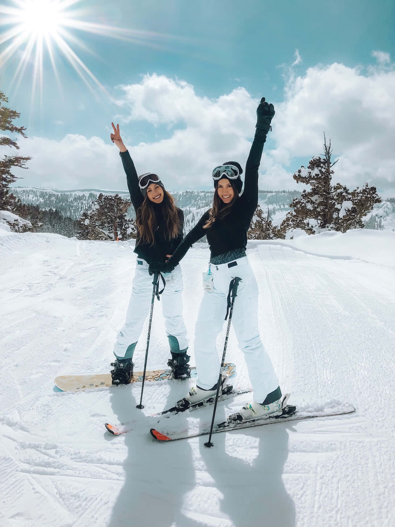 Two women in white ski pants and black ski jackets pose on the snow with their skis on - Ski