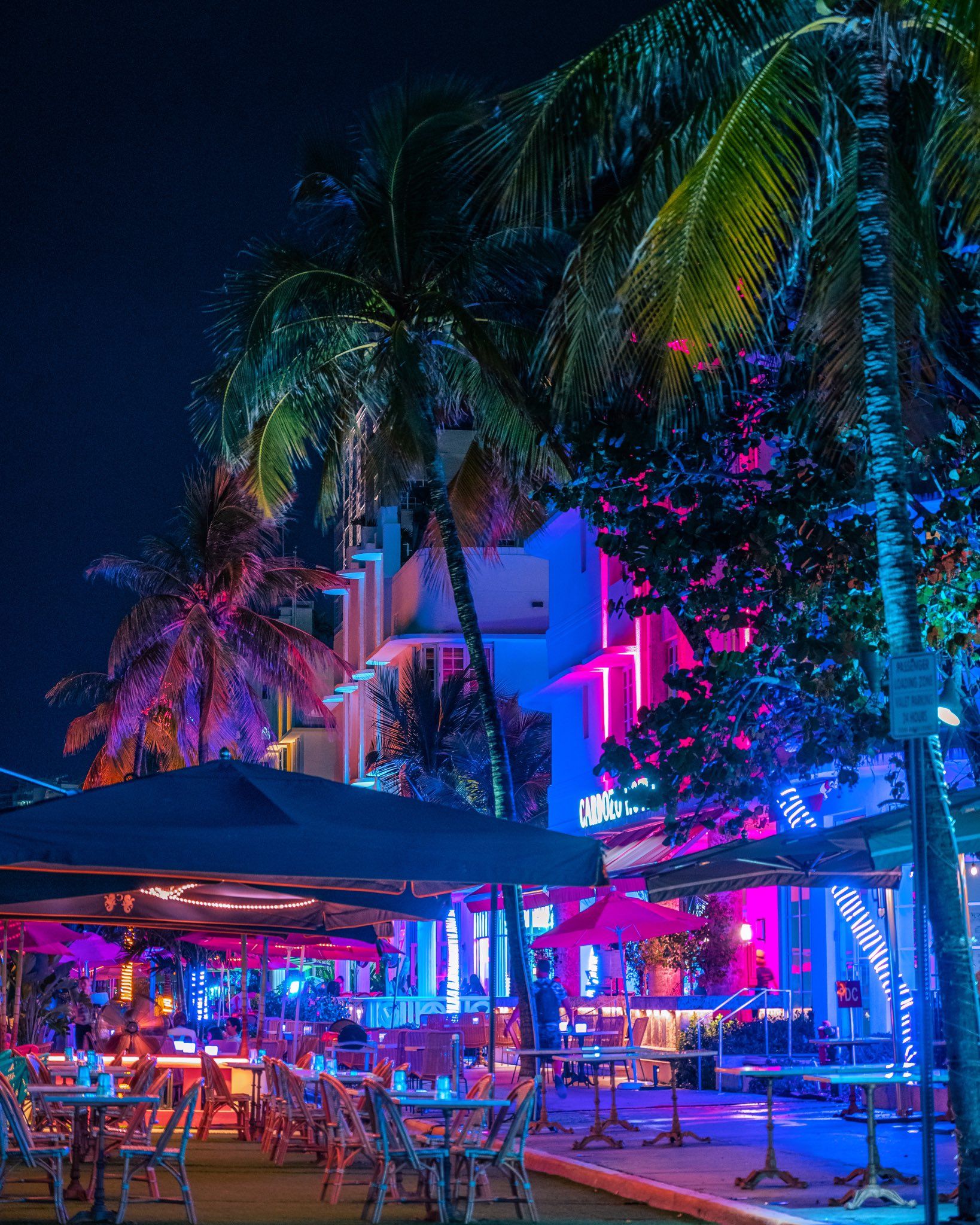 A night view of a restaurant with tables and chairs outside and neon lights on the building. - Miami