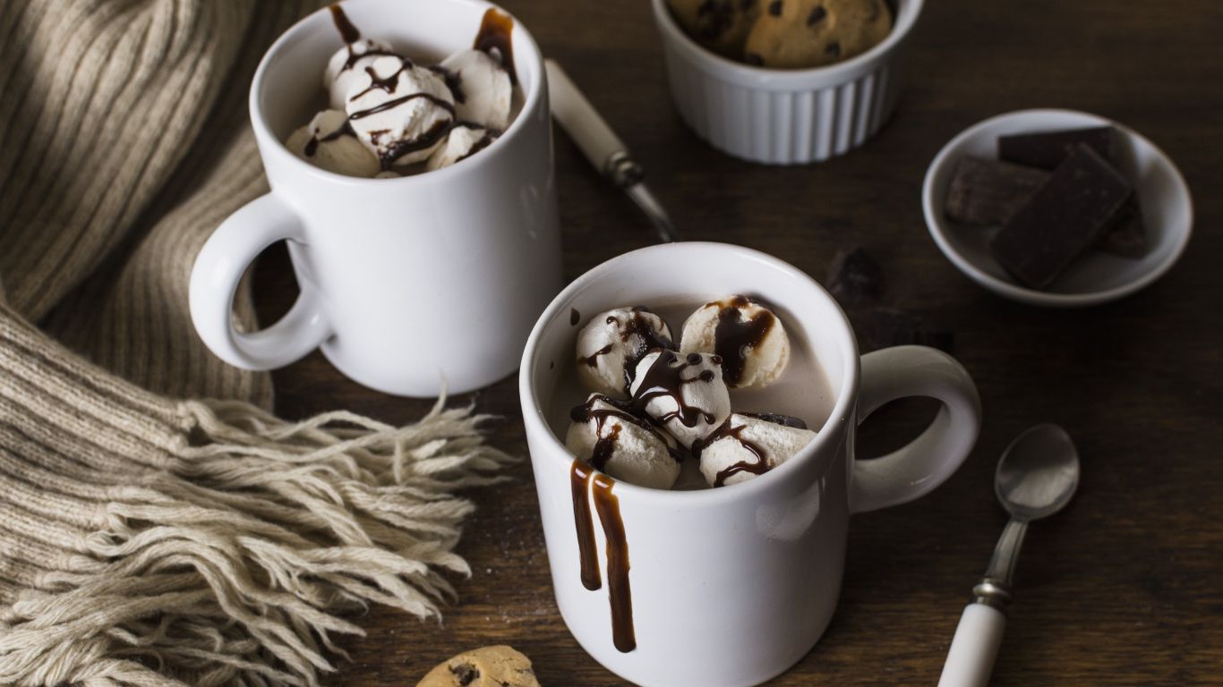 Two mugs of hot chocolate topped with marshmallows and chocolate sauce, with a blanket and cookies on the side. - Chocolate, marshmallows