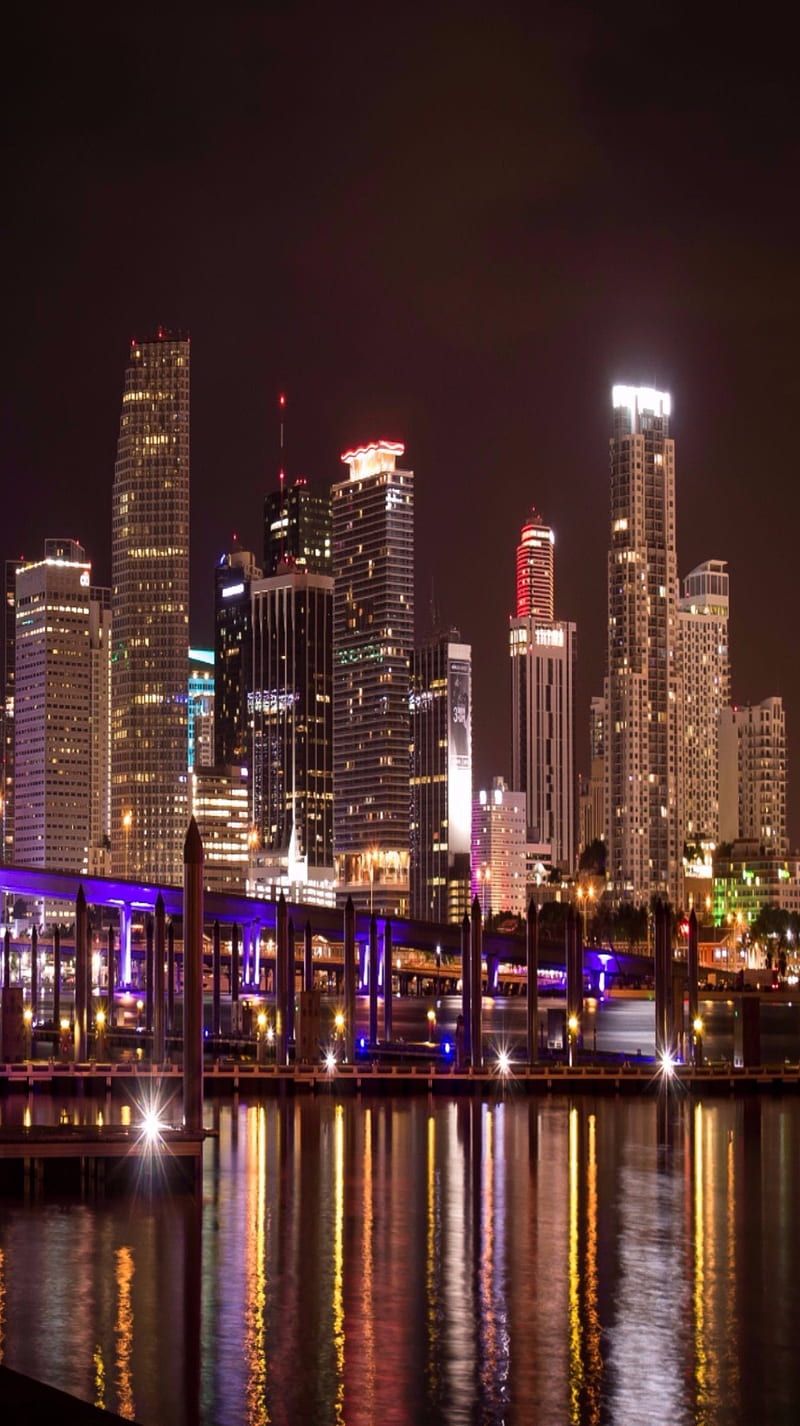 A city skyline is lit up at night with the lights reflecting off the water. - Miami