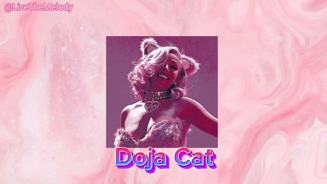 A woman with pink hair and a cat ears headband is smiling and looking to the side. The background is a pink marble. - Doja Cat