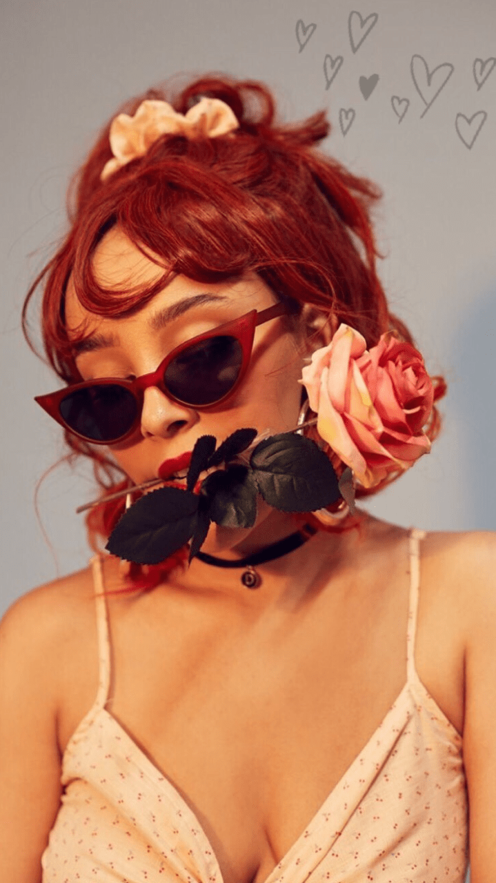 A woman with red hair and sunglasses holding a rose to her face. - Doja Cat