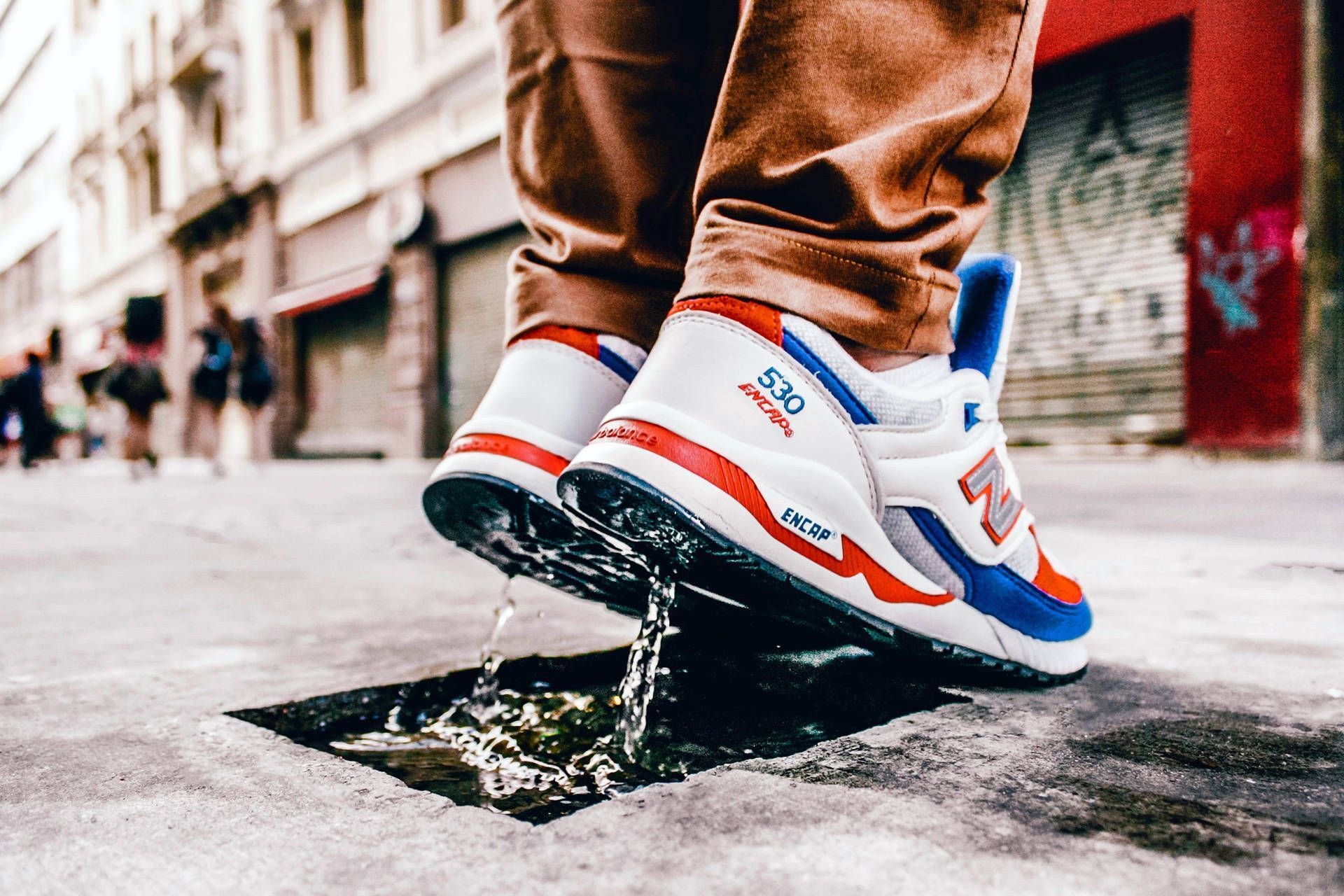 A person wearing a white sneaker with red and blue accents is stepping on a puddle. - New Balance, shoes