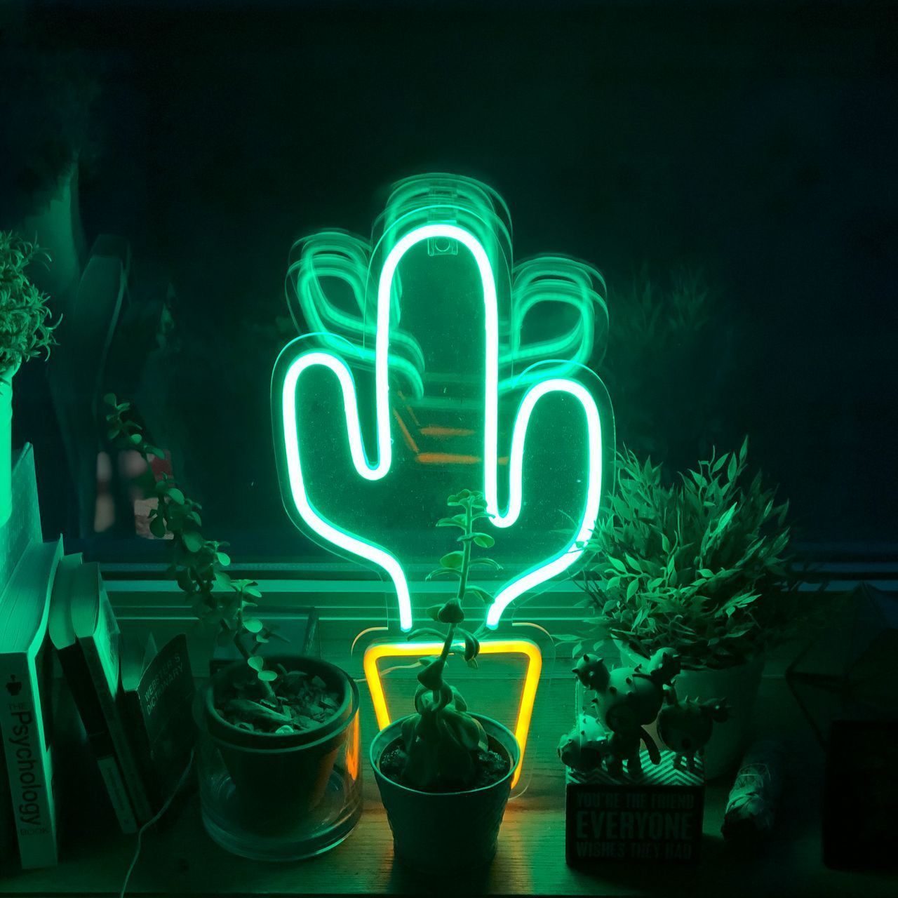 A neon cactus sign in a dark room surrounded by potted plants. - Lime green