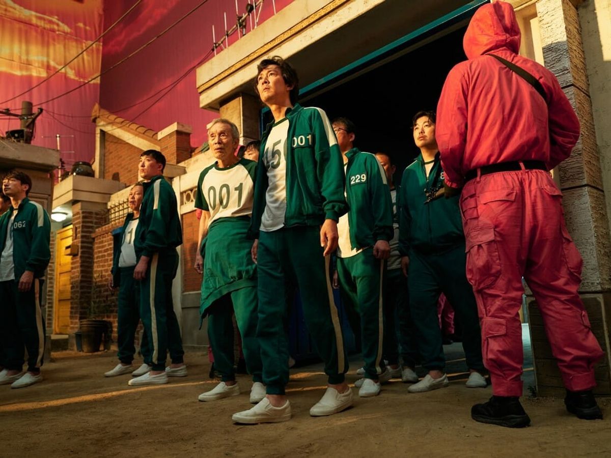 Several men in green prison uniforms stand in a line in front of a building. - Squid Game