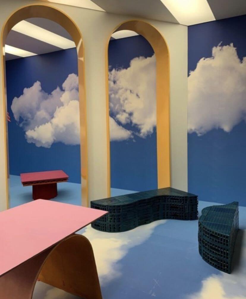 This is a photograph of a room with blue floors, white columns, and blue walls with white clouds painted on them. There are three cloud-shaped benches and a pink table. - Weirdcore