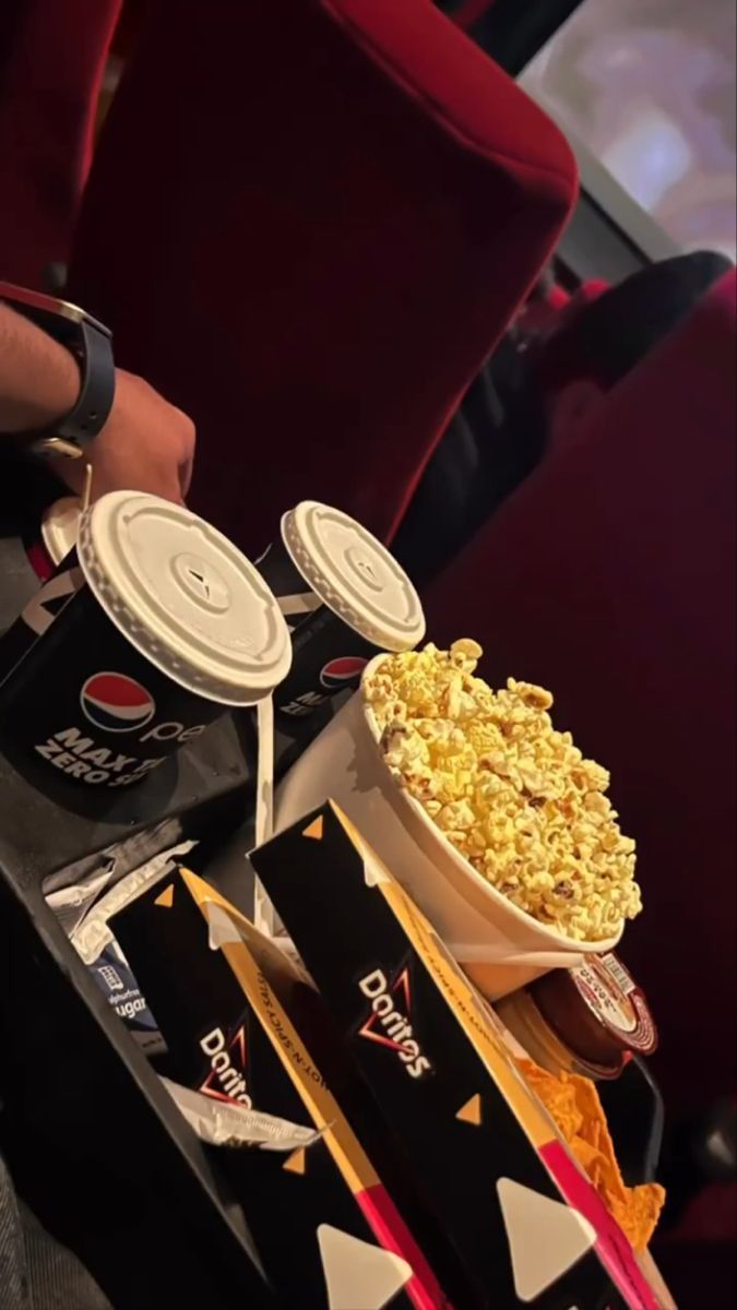 A bowl of popcorn and a cup of soda sit on a chair in a movie theater. - Doritos