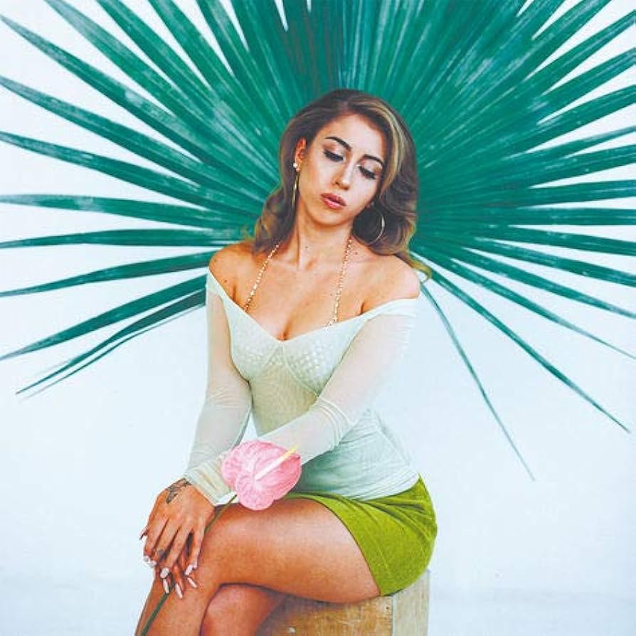 A woman sitting on a stool with a green palm frond behind her - Kali Uchis