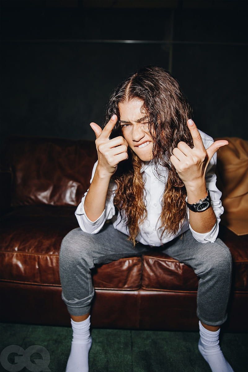 A young man with long hair sitting on a couch making a rock sign with his fingers - Zendaya