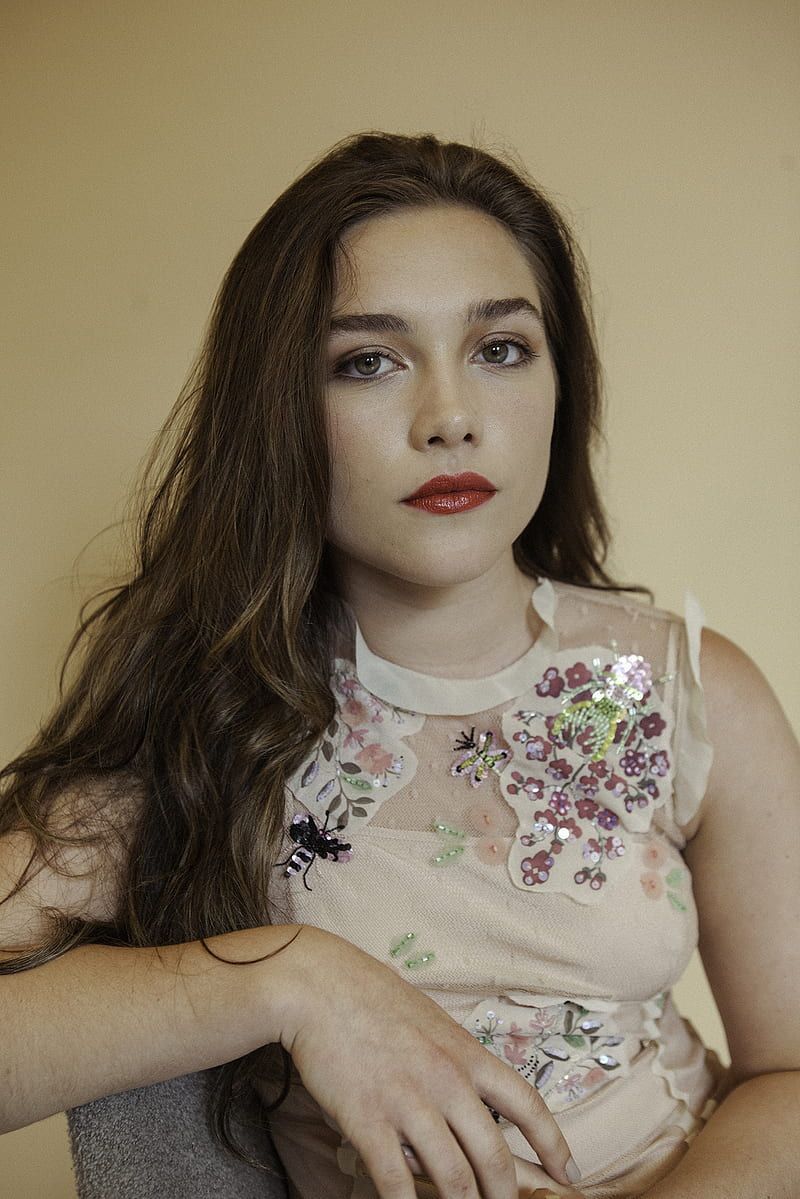 A woman with long brown hair and red lipstick sits on a chair. - Florence Pugh